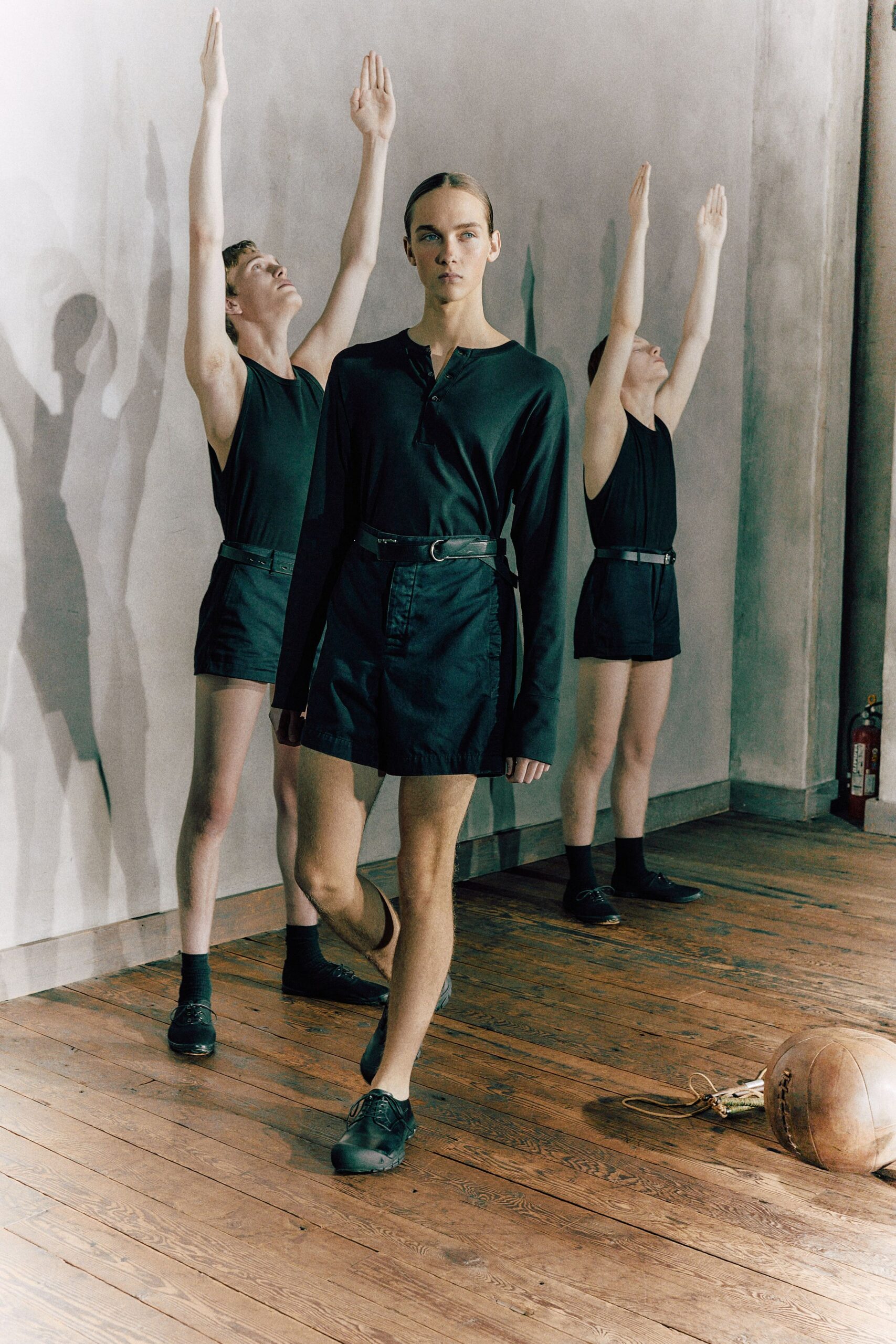 Black jersey henley, shallow placket left casually half-unbuttoned. High-waisted black shorts in crisp cotton twill. Slim leather belt with a dull silver D-ring. Black leather derbies, plain and almost school-uniform stark. After drills in a drafty gym, medicine ball abandoned by the wall.