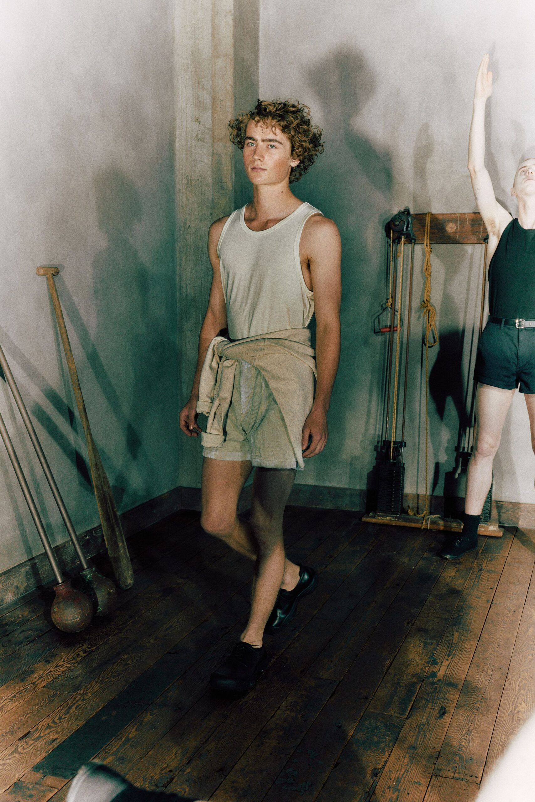 Off-white cotton jersey tank with deep armholes cut almost to the waist. Pale sweatshirt knotted low around the hips, sleeves hanging unevenly. Rolled light shorts in soft knit, hem peeking below the tied layer. Black slip-on shoes with blunt rounded toes. For the quiet stretch between sets in a wood-floored gymnasium.