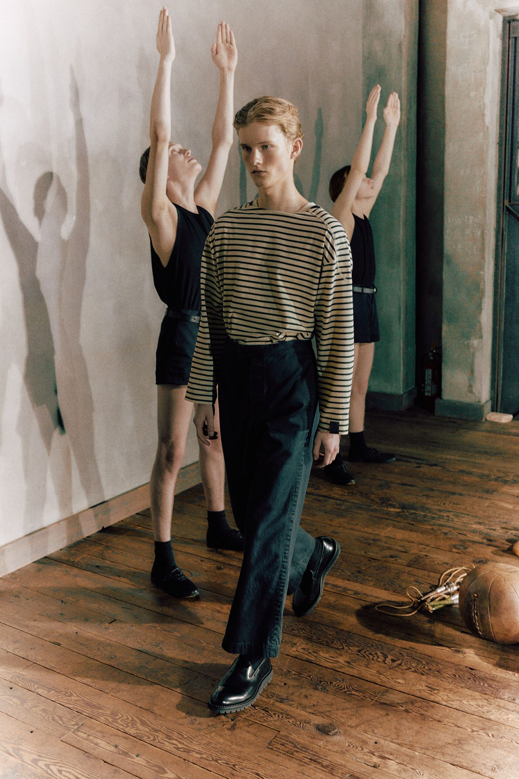 Cream jersey boatneck with narrow black sailor stripes and dropped shoulders. Inky high-waisted trousers, pressed flat with a slight crop. Black leather loafers with thick treaded soles. Walking past the gym mats after morning movement class lets out.