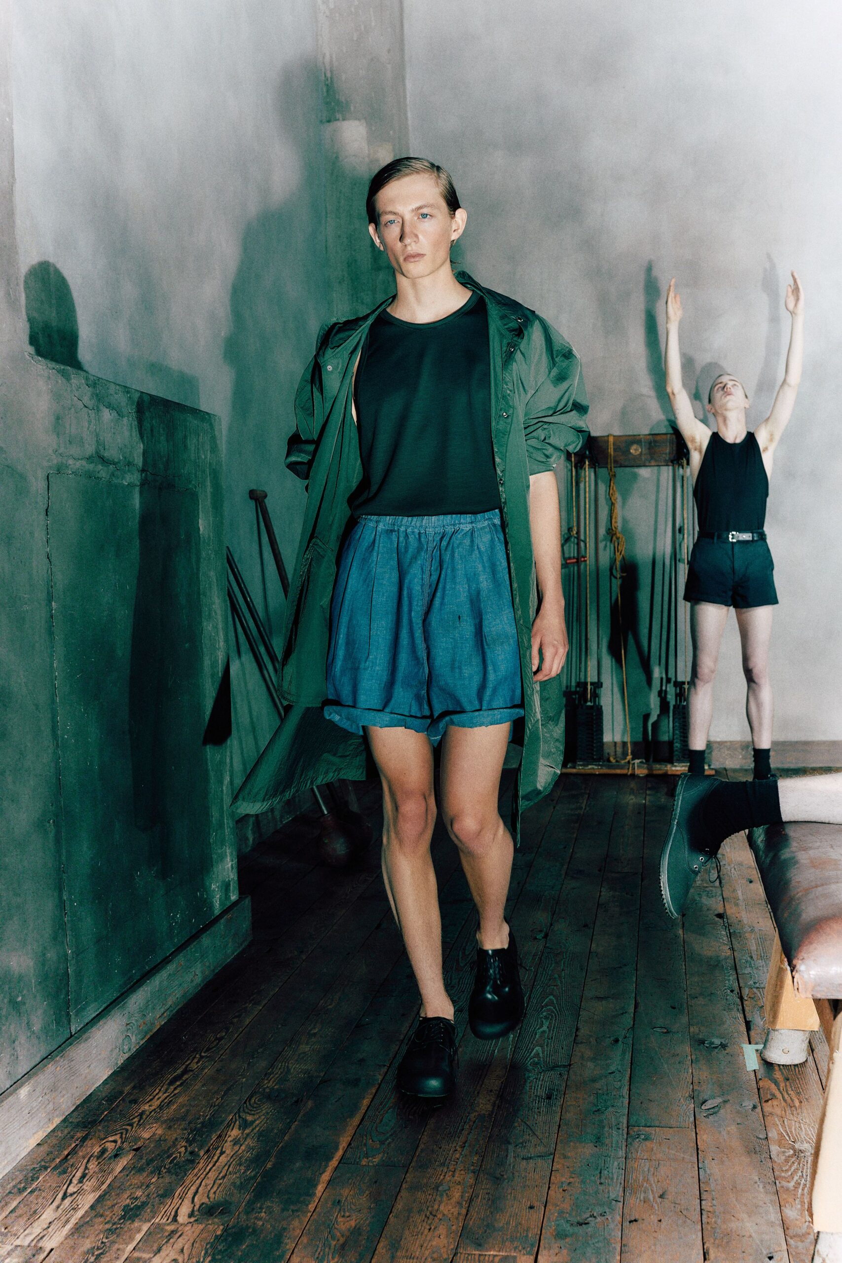 Forest nylon parka, snap placket hanging open and hem kicking sideways. Black ribbed sleeveless top cut loose through the armhole. Chambray shorts with deep pleats and a softly gathered waistband. Black leather lace-ups with bulbous rounded toes. Walking across old floorboards in a drafty house before the fire catches.