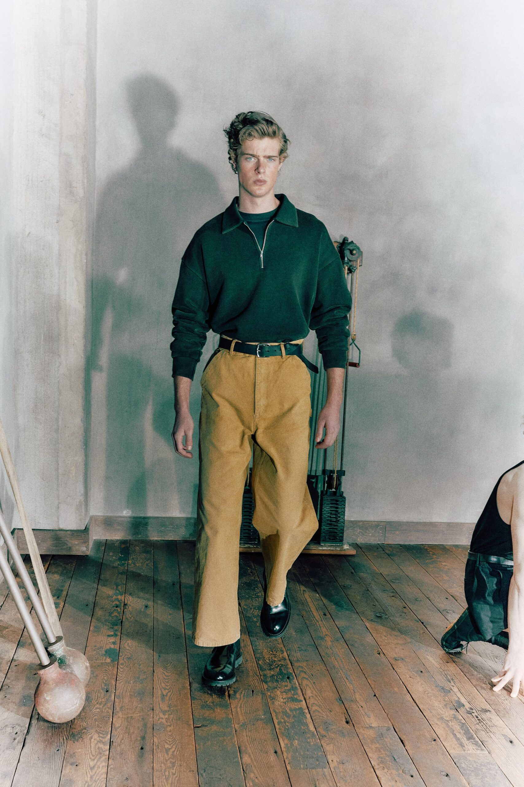Deep pine knit quarter-zip, collar spread wide above a crewneck layer. Mustard corduroy trousers, pressed crease falling from a high belted waist. Black leather belt, strap tail jutting past the buckle. Black leather shoes with a chunky lug sole. Walking across old floorboards in a drafty studio before the heaters catch.