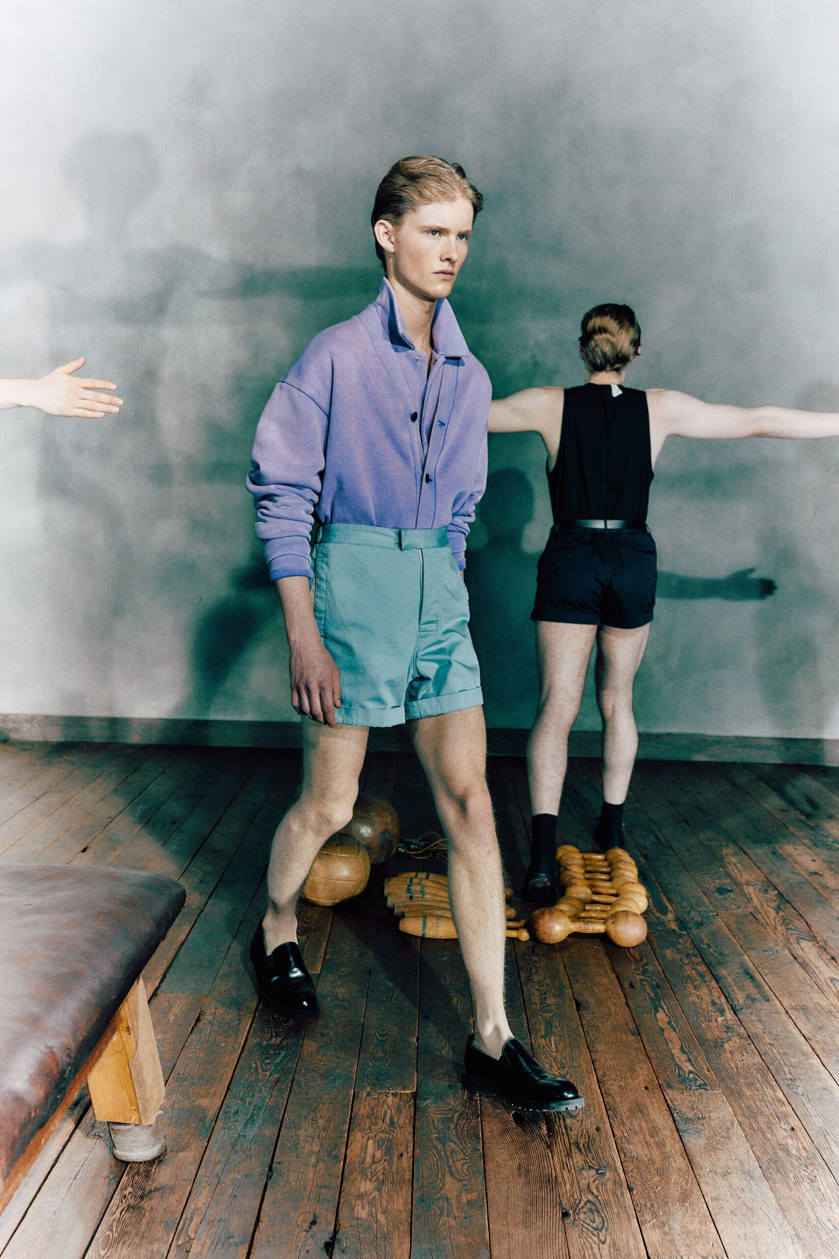 Periwinkle fine-knit cardigan, collar flipped high and cuffs pushed dense at the wrist. Powder-blue tailored shorts with a deep cuff and tabbed waistband. Black leather penny loafers with thick lug soles. Walking across old gym floorboards after somebody dragged the apparatus out.