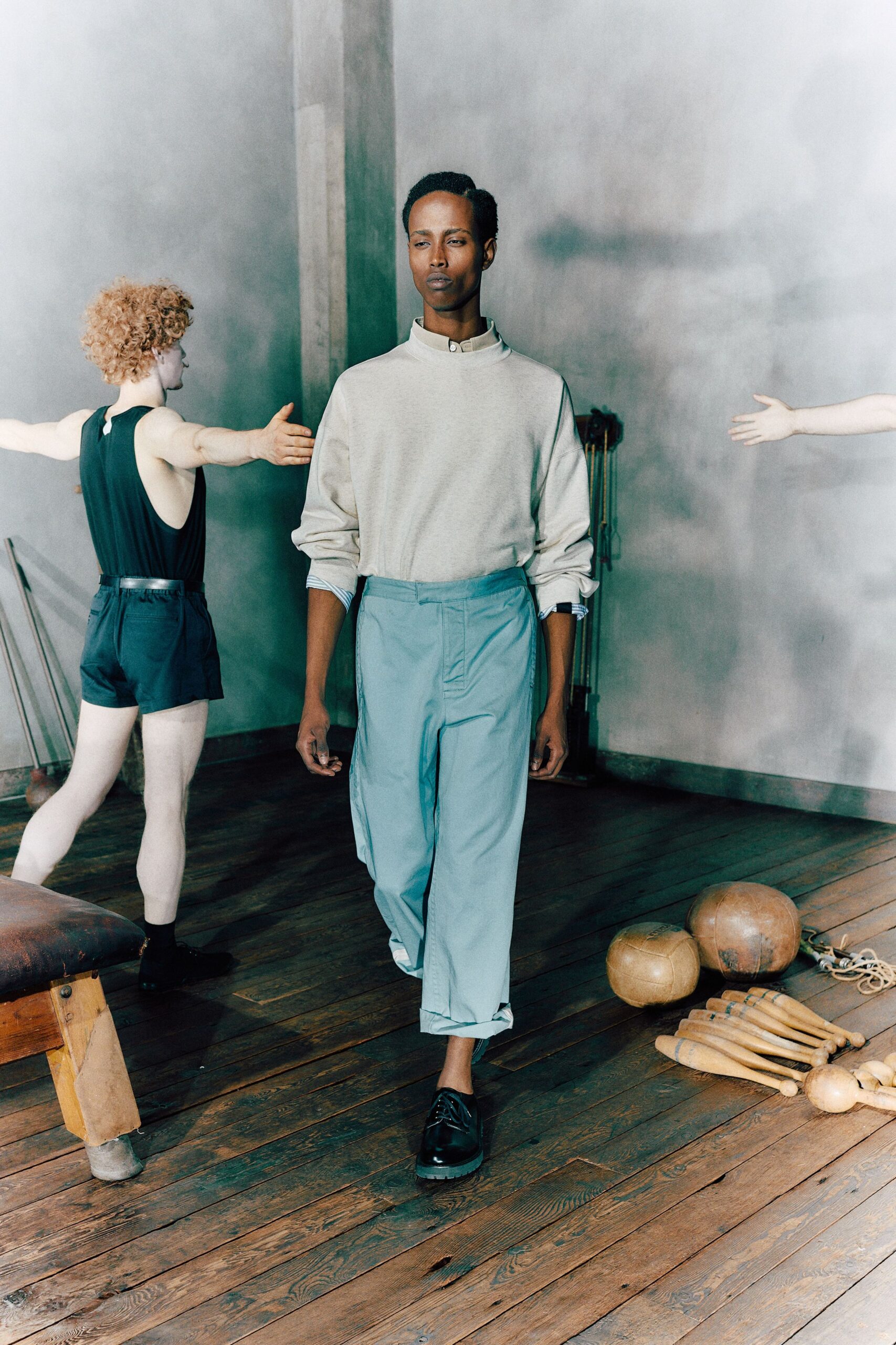 Pale stand-collar shirt peeking above the neck with one top button. Bone fine-knit sweater with dropped shoulders and sleeves pushed bluntly up. Powder-blue cropped trousers, pressed flat and rolled once at the hem. Black derbies with thick rubber soles. Walking past medicine balls and wooden clubs in a half-lit gymnasium.