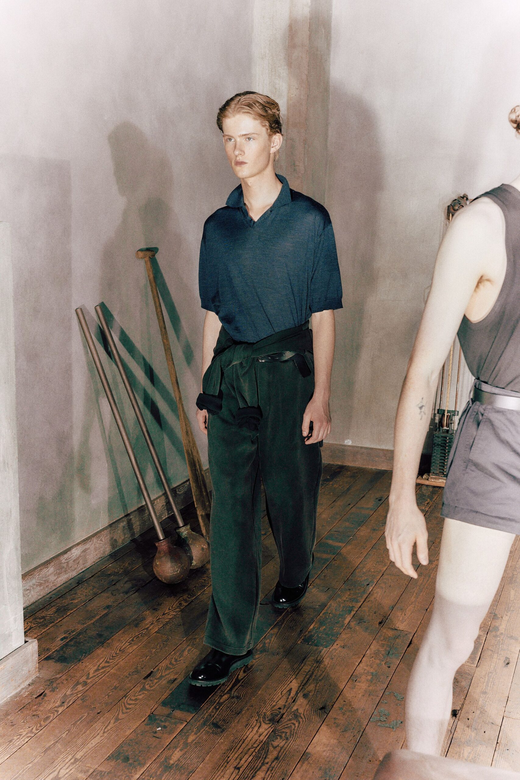 Inky fine-knit polo, open collar standing slightly away from the throat. Dark wide-leg trousers with a soft drape and pressed front crease. Second knit tied low at the waist, sleeves knotted off-center. Black leather shoes with bulbous toes and thick soles. Walking through a drafty house while the floor varnish is still drying.