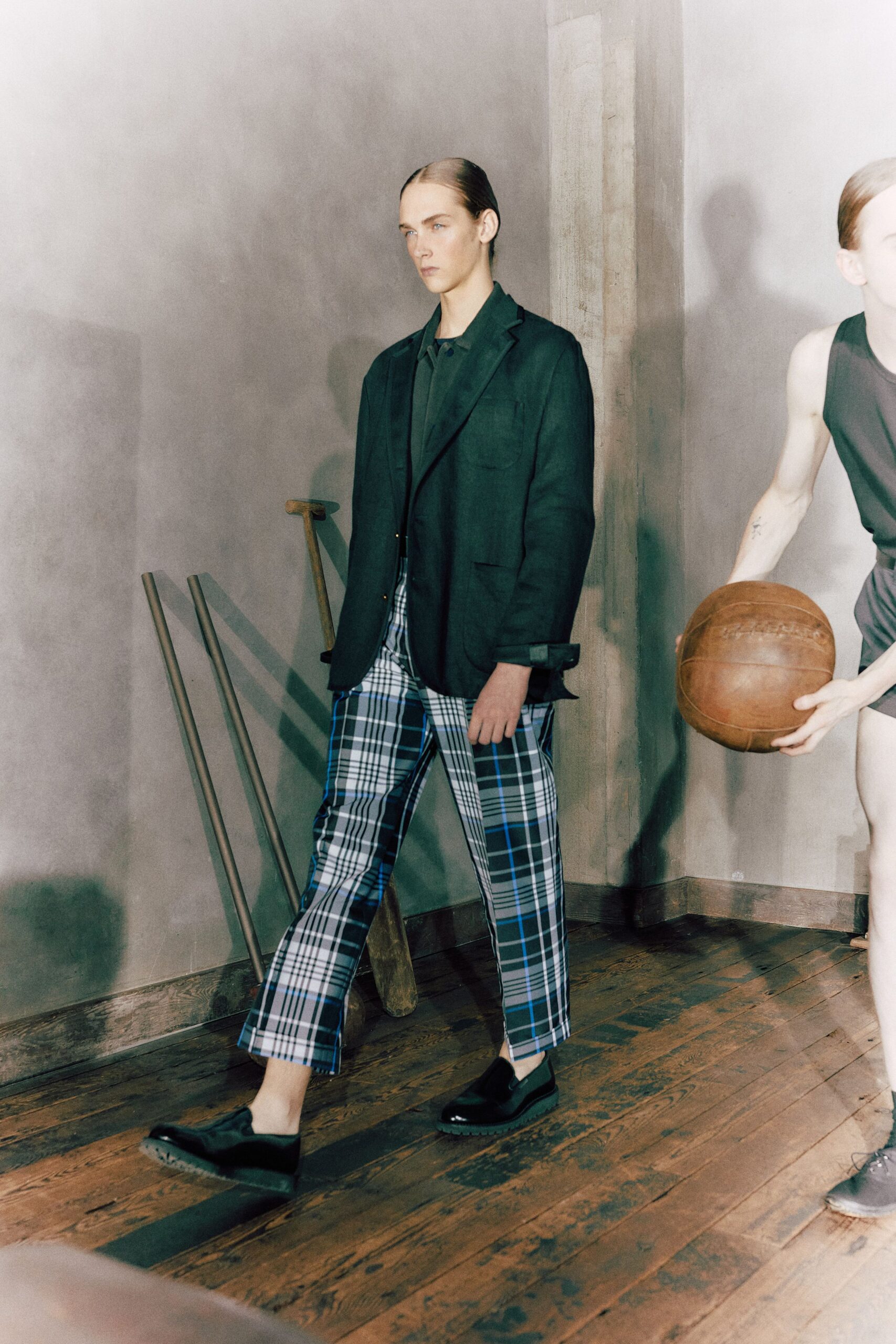Dark forest blazer in soft matte suiting, cuffs turned back once. Matching button-front shirt with a small flat collar. Cropped plaid trousers streaked with cobalt lines through the check. Black leather slip-ons with thick ridged soles. Crossing a gym floor after hours while the old equipment stays stacked by the wall.