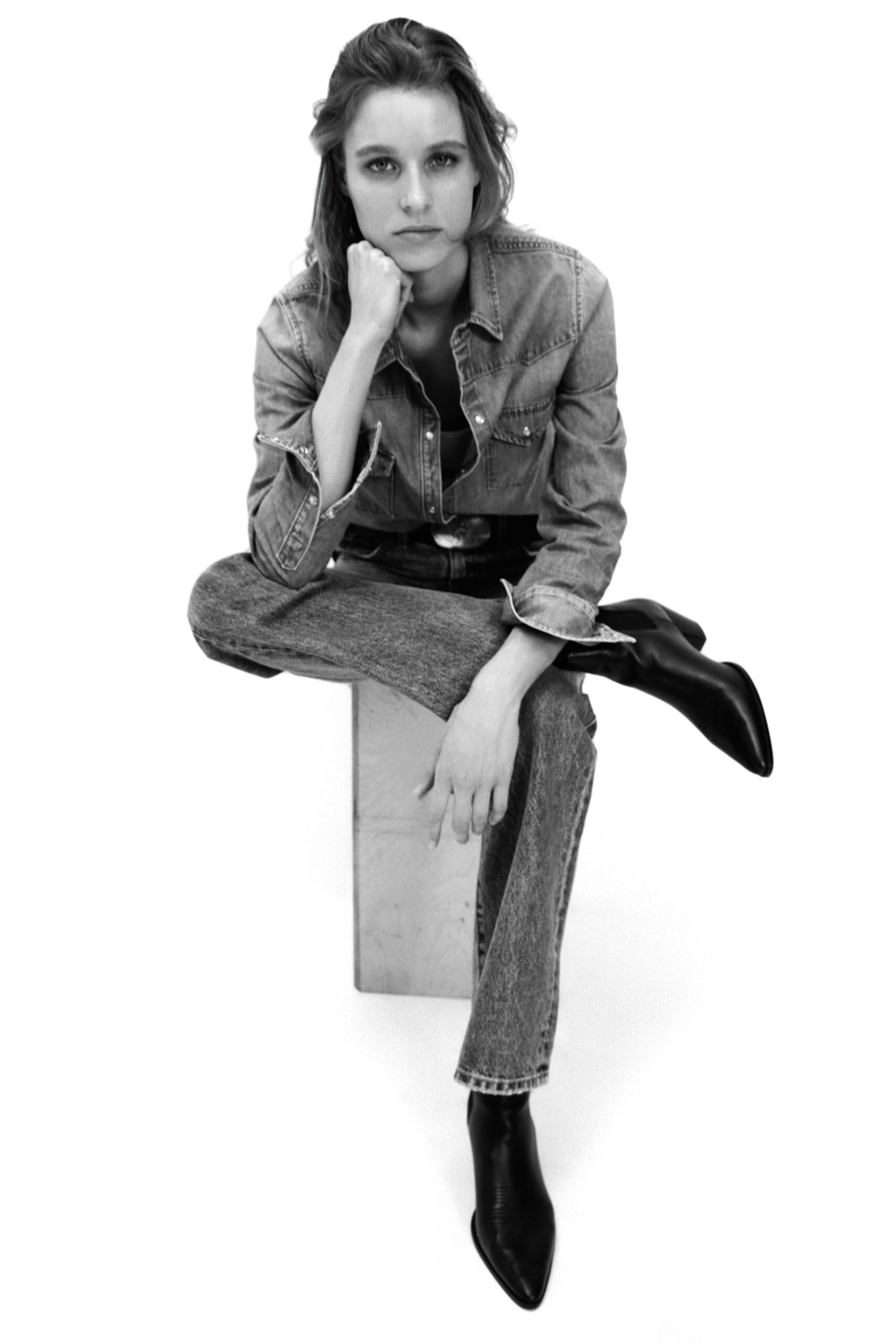 Faded denim shirt with pearly snaps and one cuff turned back. Wide leather belt with a bright oval buckle. Straight acid-wash jeans, cropped just above the ankle. Black leather ankle boots with narrow pointed toes. Sitting on a studio plinth while the freight elevator sticks again.