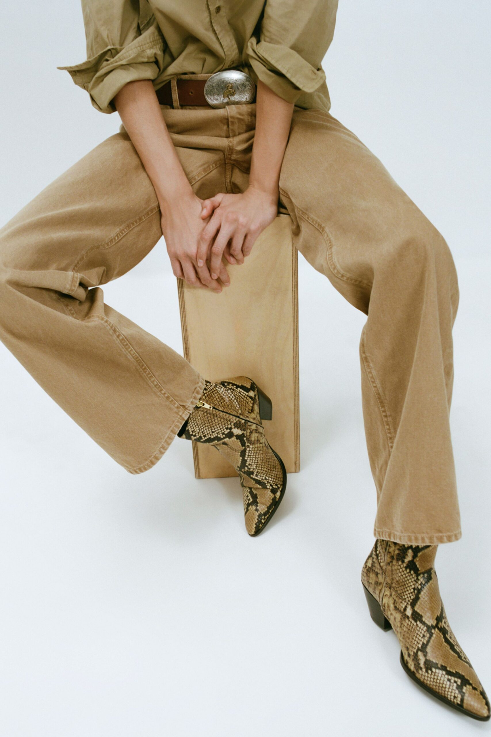 Khaki cotton shirt with sleeves pushed into thick rumpled cuffs. Brown leather belt with an oversized engraved silver buckle. Sandwashed tan jeans, wide through the leg with turned hems. Python ankle boots with pointed toes and stacked block heels. Sitting on a feed crate while the tack room finally cools down.