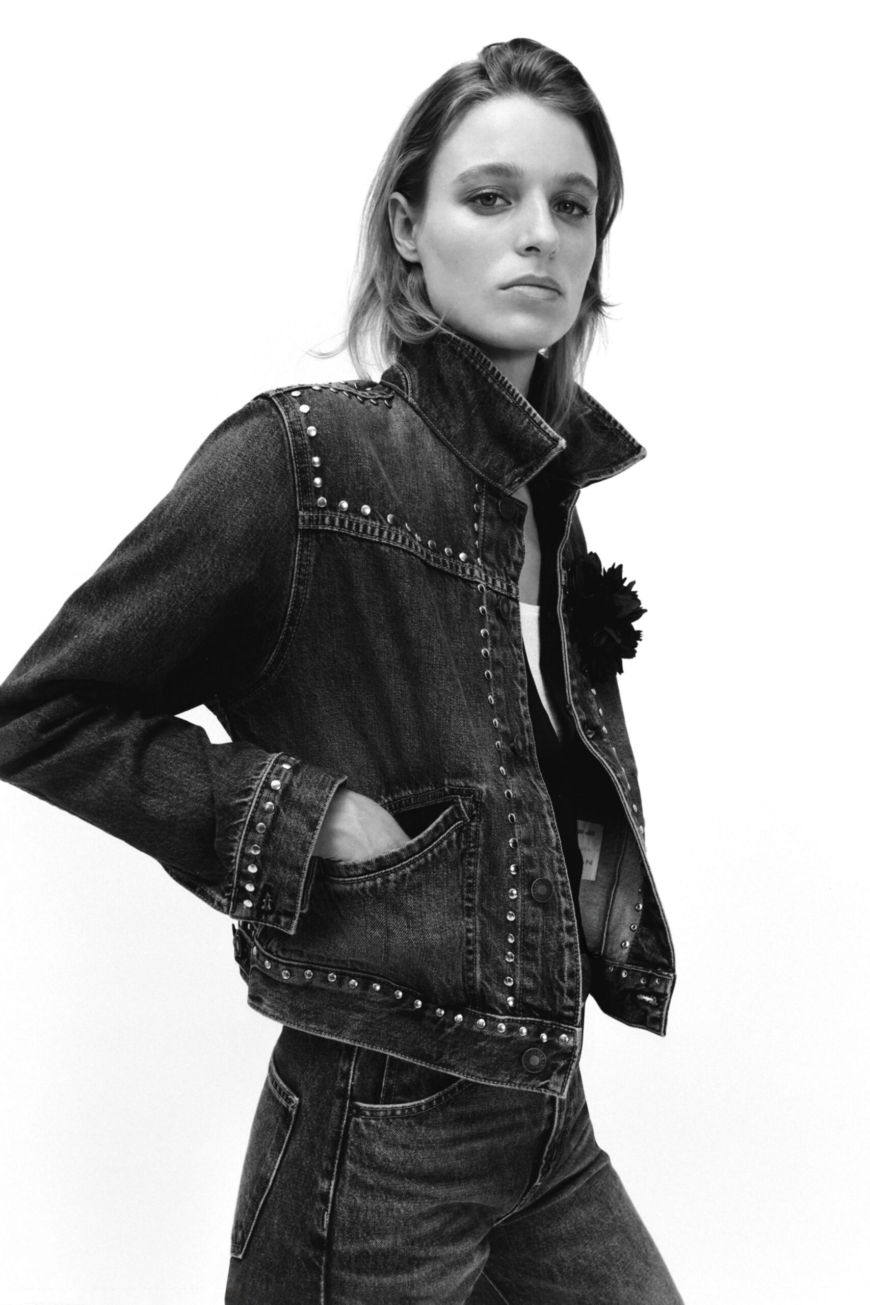Black-wash denim jacket, stand collar flipped high and seams traced in silver studs. White scoop-neck top just visible beneath the placket. Black fabric bloom pinned off-center at the chest. Matching dark denim jeans with a long coin-pocket stitch line. For buying cigarettes at a roadside bar before the rain starts.