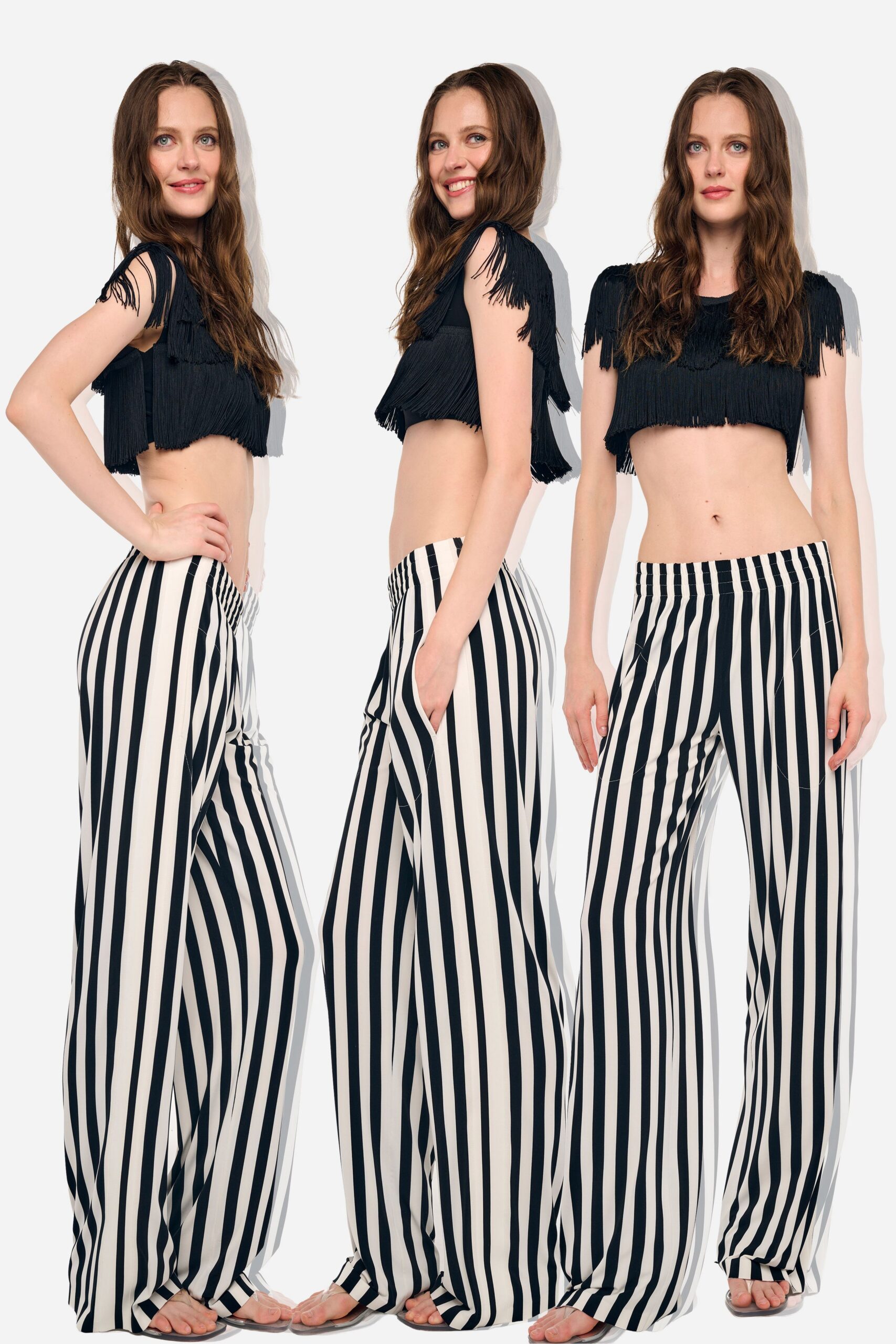 Black cropped top with dense fringe tiers and shoulder tassels. Cream palazzo trousers in stark black vertical stripes, pooling at the foot. Clear flat sandals. Walking the boardwalk arcade before the heat breaks.