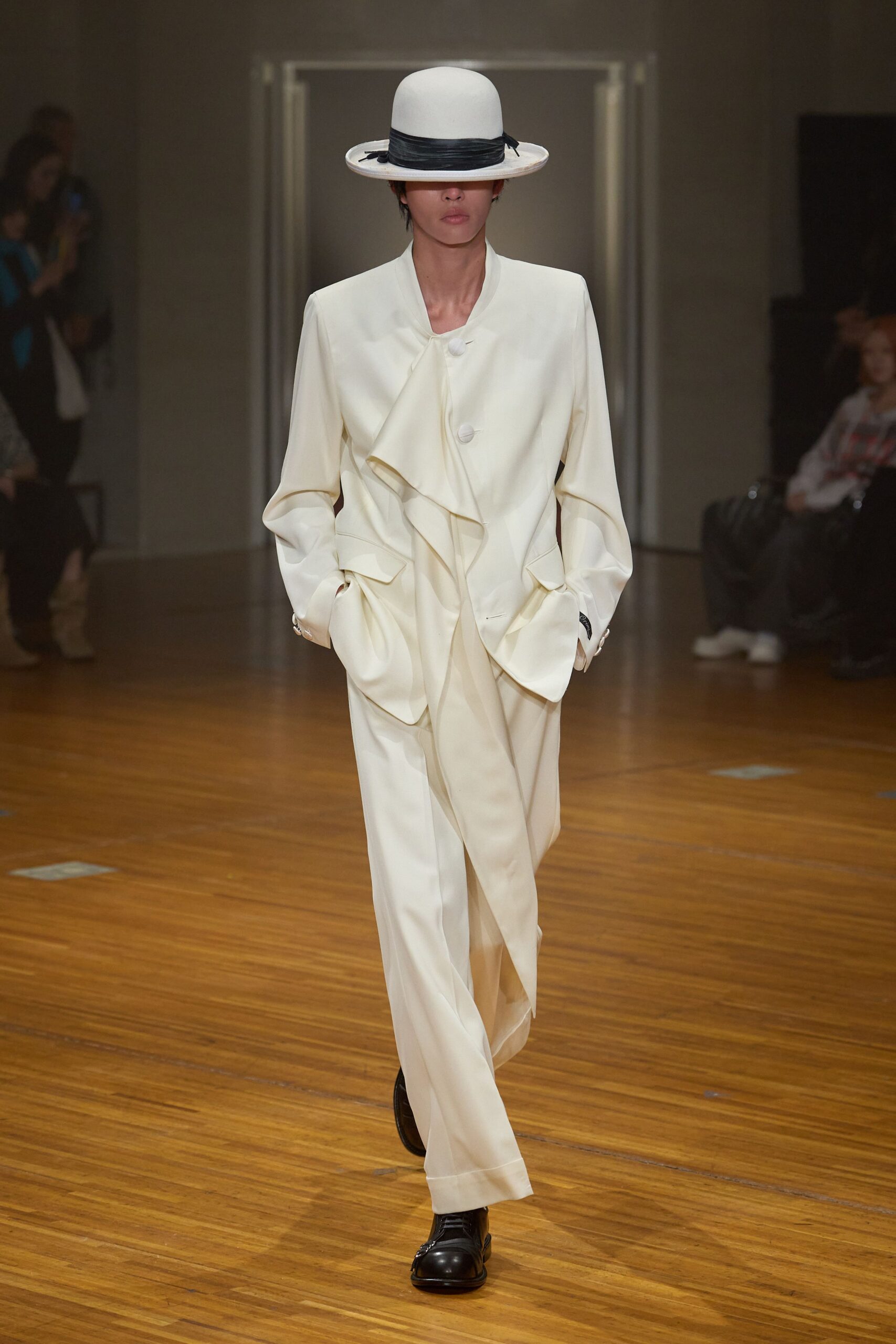White felt boater with a black grosgrain band dropping over the eyes. Ivory jacket cut with a cascading front ruffle and covered buttons. Wide ivory trousers with a soft break and deep turned cuffs. Black leather lace-ups with a rounded toe. Crossing a parquet ballroom at noon before the wedding guests arrive.