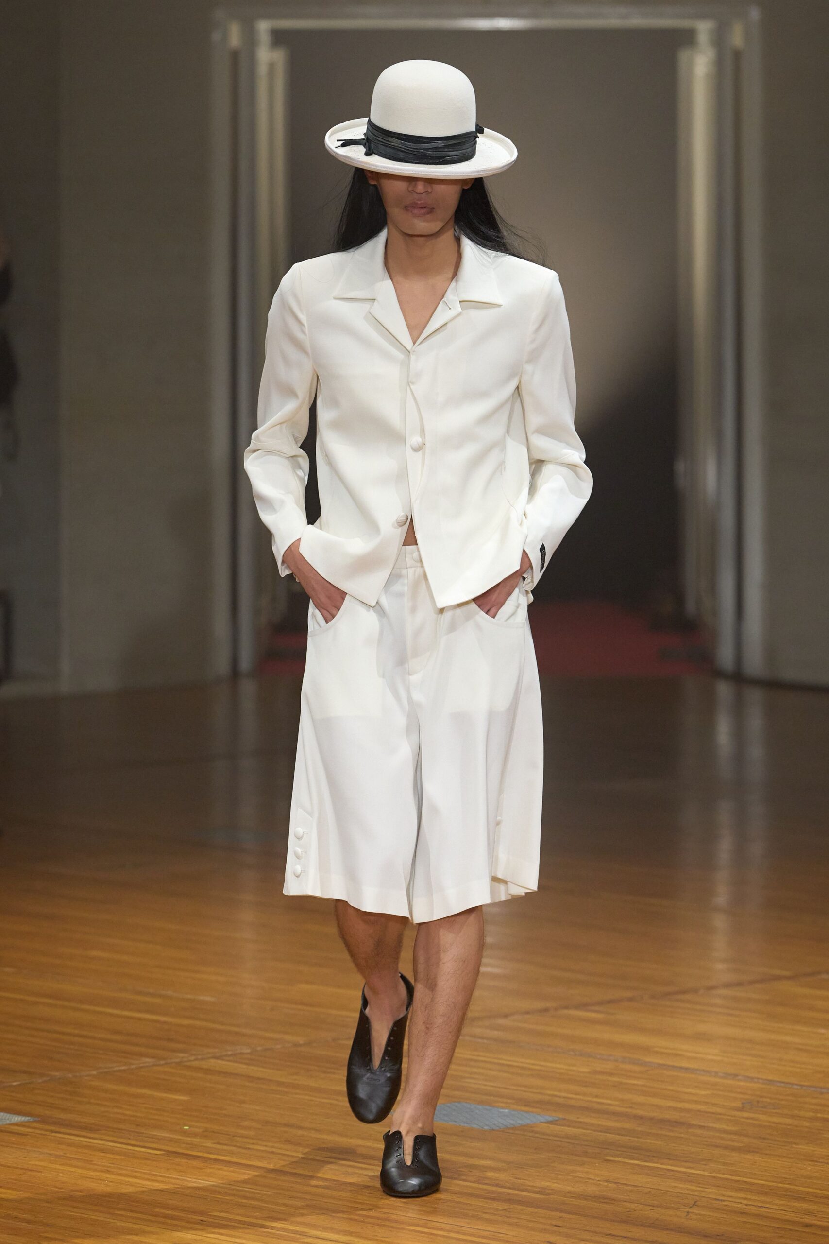 White felt bowler with a black wrapped band, brim tipped low. Ivory tailored jacket with broad notch lapels and covered buttons. Matching calf-length culottes, pressed wide with three buttons at the side hem. Black leather lace-ups cut with a narrow split vamp. Crossing a museum parquet floor before the public is let in.