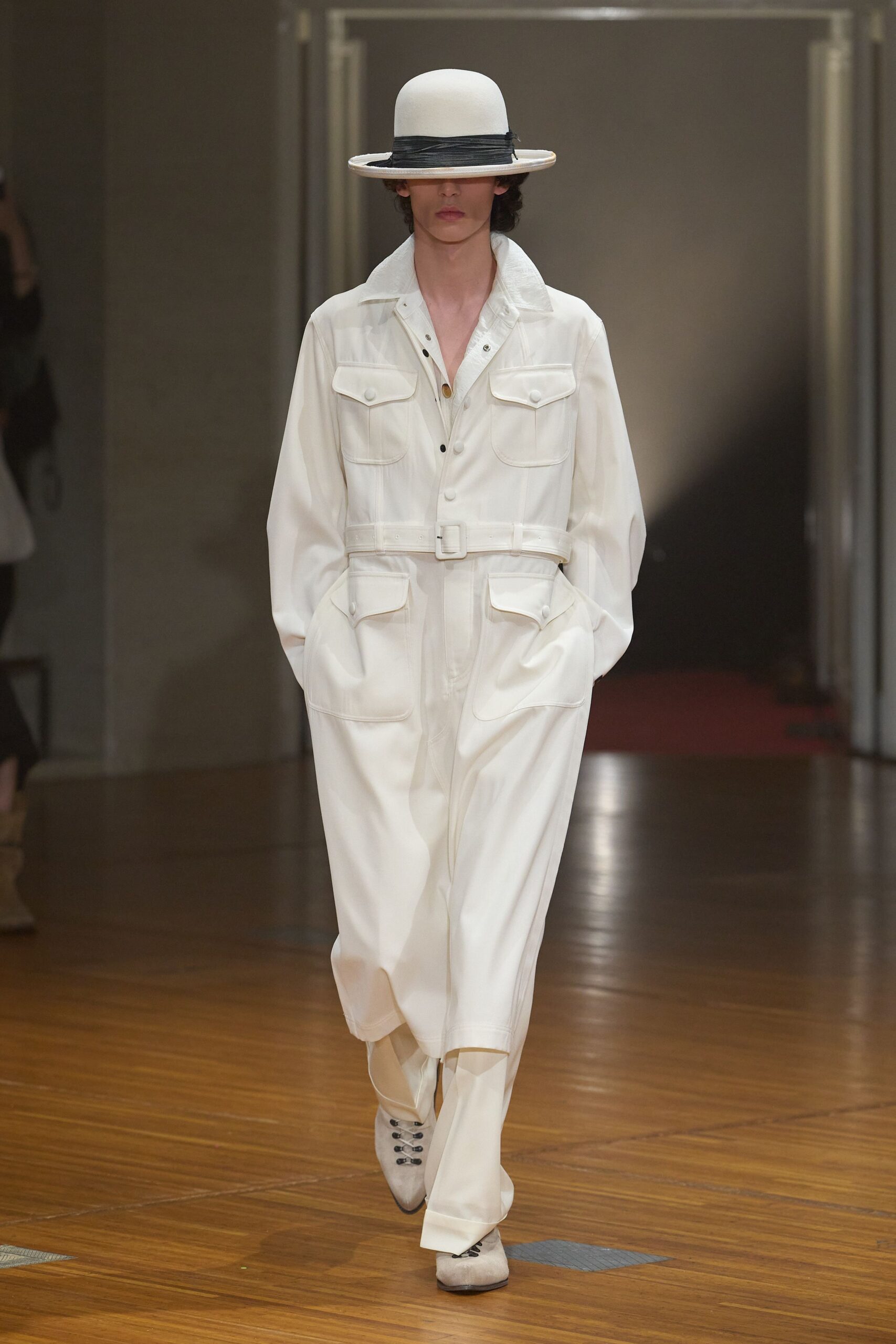 White bowler with a low brim and narrow black wrapped band. Milk-white utility jumpsuit, belted waist and four bulbous flap pockets. Pointed pale suede boots laced with five black hooks. Crossing a near-empty station concourse just after the floors were buffed.