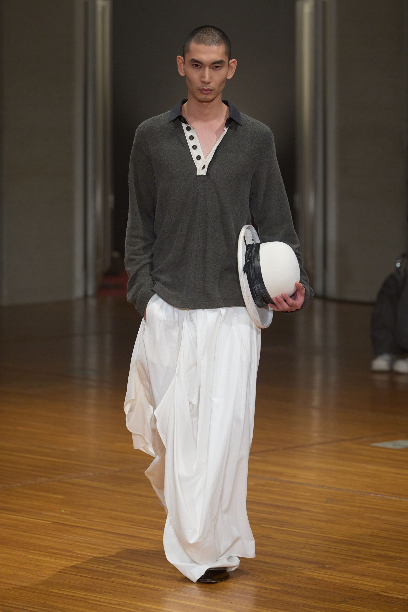 Charcoal boucle polo shirt with a white placket and dark collar. Milk-white trousers cut extremely wide, pooling into soft vertical folds. White hard-shell hat carried with a black wrapped band. Walking across a ferry deck before the wind fully picks up.
