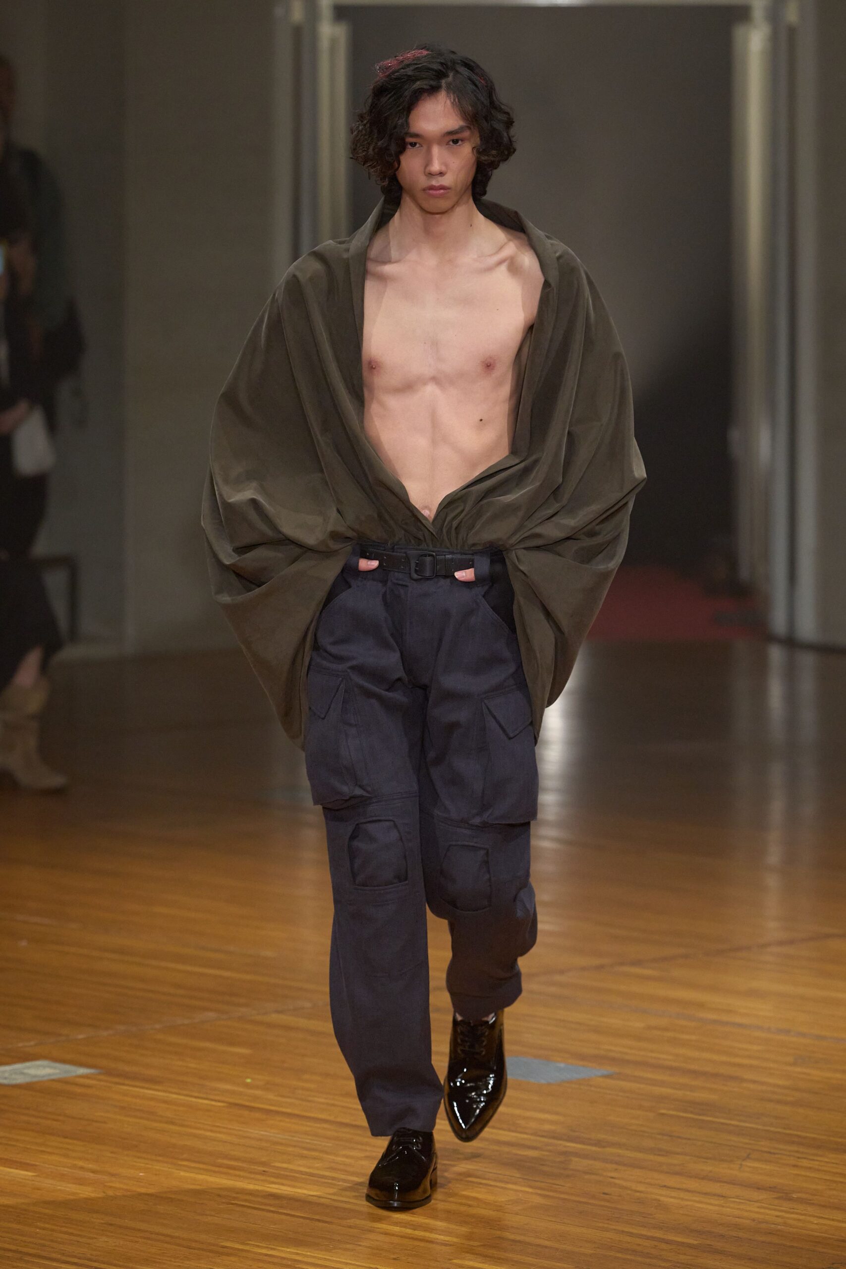 Olive blouse in crisp cloth, plunged open and ballooning around the arms. Ink cargo trousers with bulky patch pockets and a webbing belt. Black patent square-toe shoes. Walking the long hallway after midnight, shirt half-fastened from somewhere else.