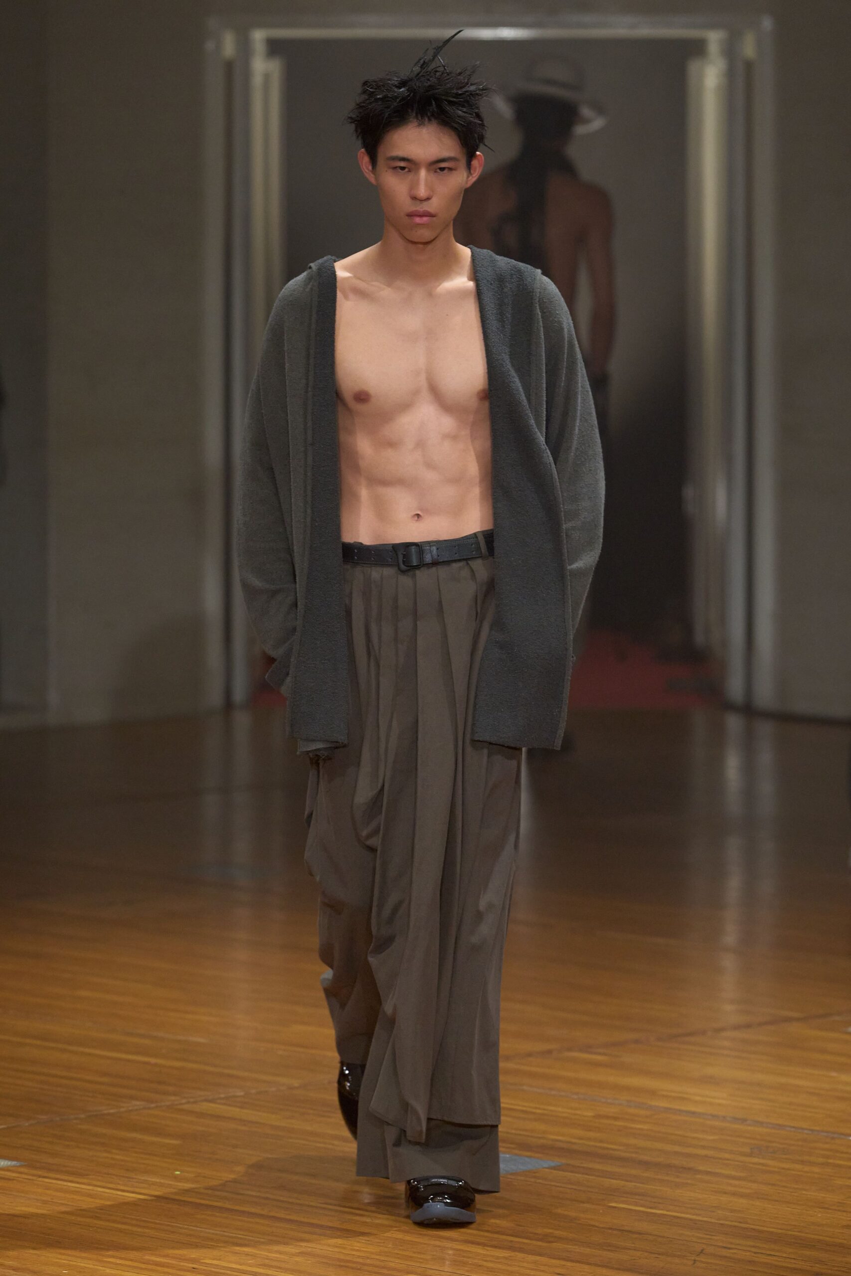 Open charcoal boucle cardigan hanging low off the shoulders. Black belt with a square buckle at the bare waist. Taupe pleated wide-leg trousers, one front panel draped loose. Black patent shoes. Crossing a museum hall after closing, still warm from the crowd.