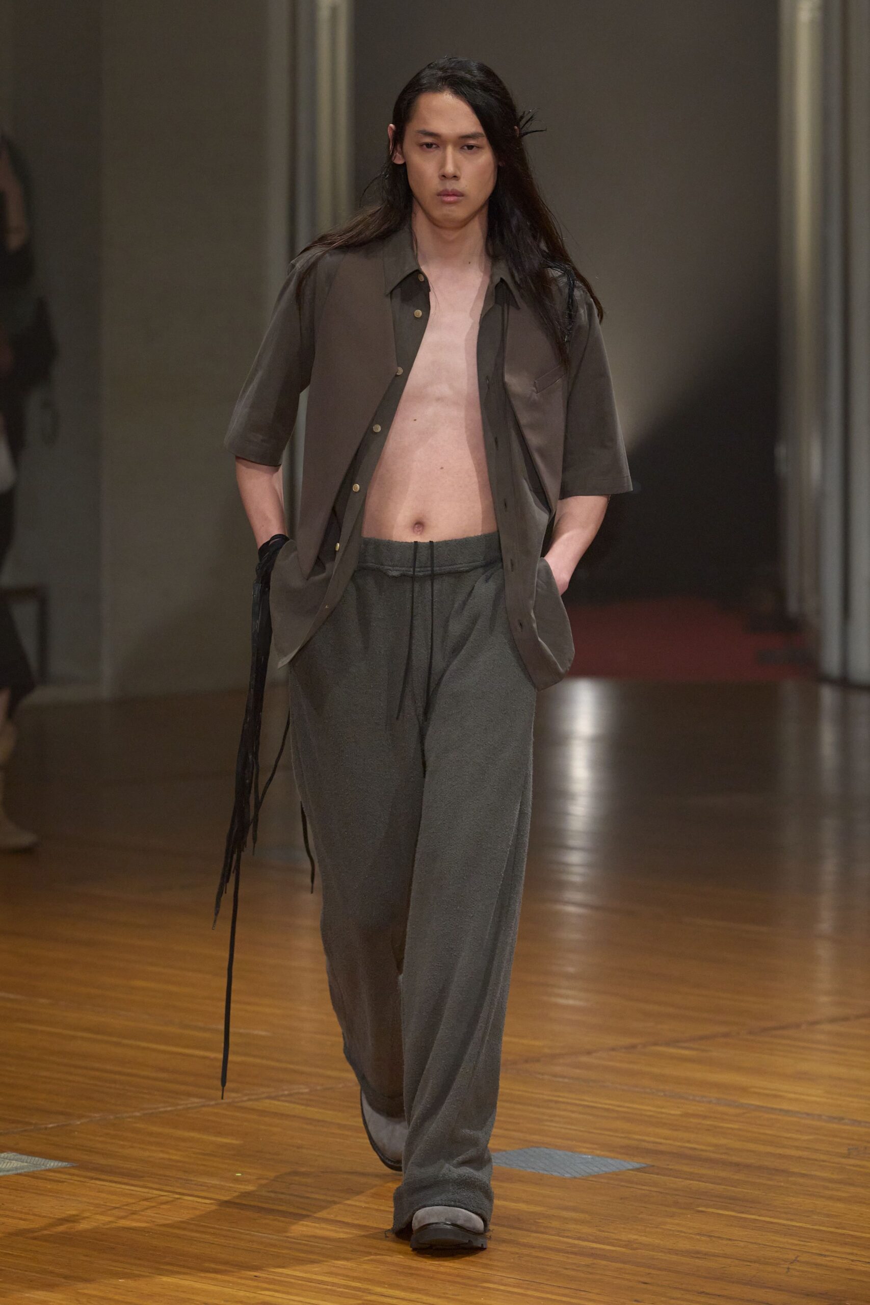 Smoke-brown short-sleeve shirt hanging open, chest pocket barely lifting the front. Charcoal loopback jersey trousers with a long black drawcord hanging center front. Square-toed dark shoes with pale felted uppers peeking out. For crossing a quiet hotel corridor before anyone else is awake.