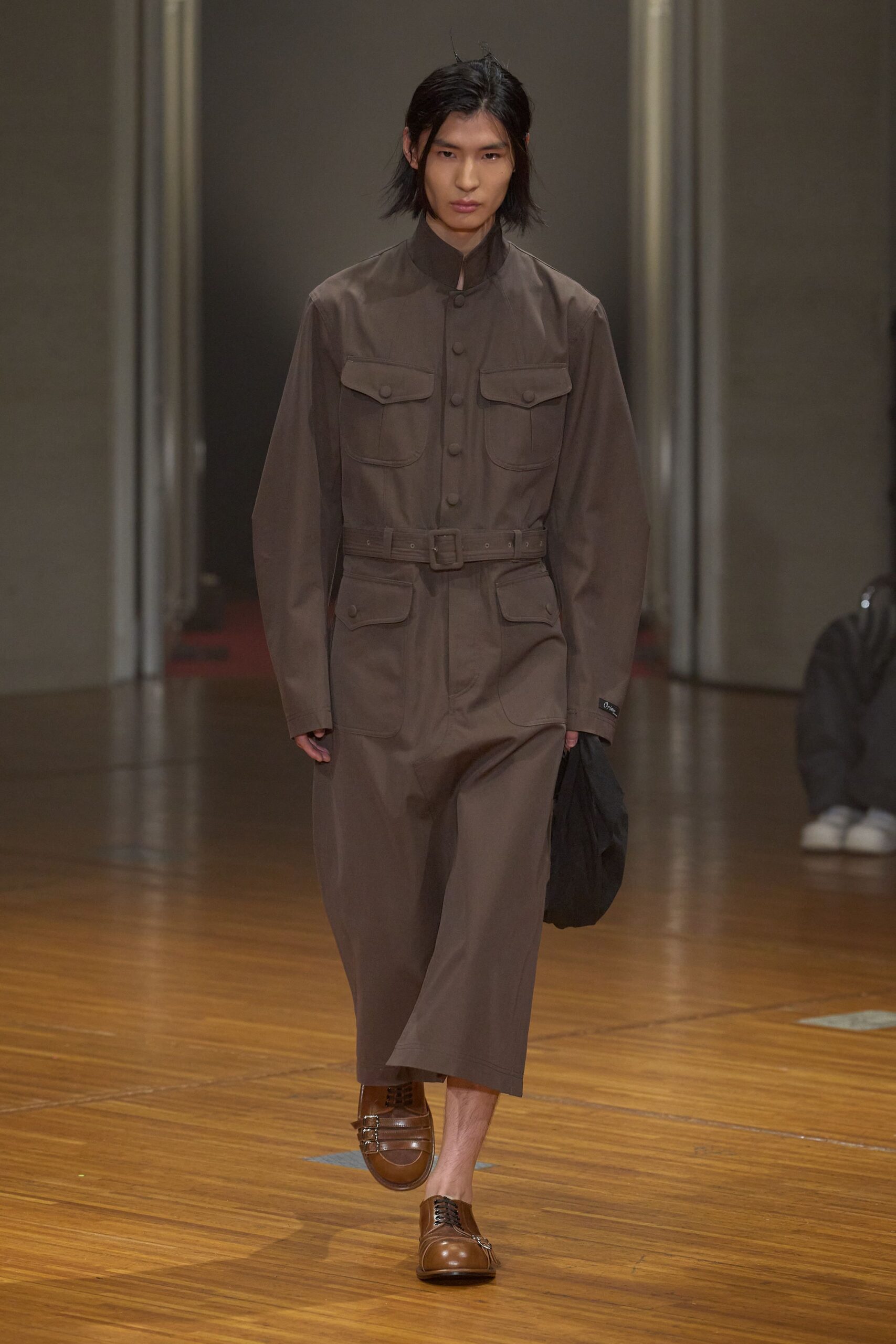 Taupe twill jumpsuit with a stand collar, covered buttons and belted waist. Soft black pouch crushed in one hand. Brown leather loafers with silver monk straps at the side. For crossing a drafty municipal hall with papers nobody else finished.