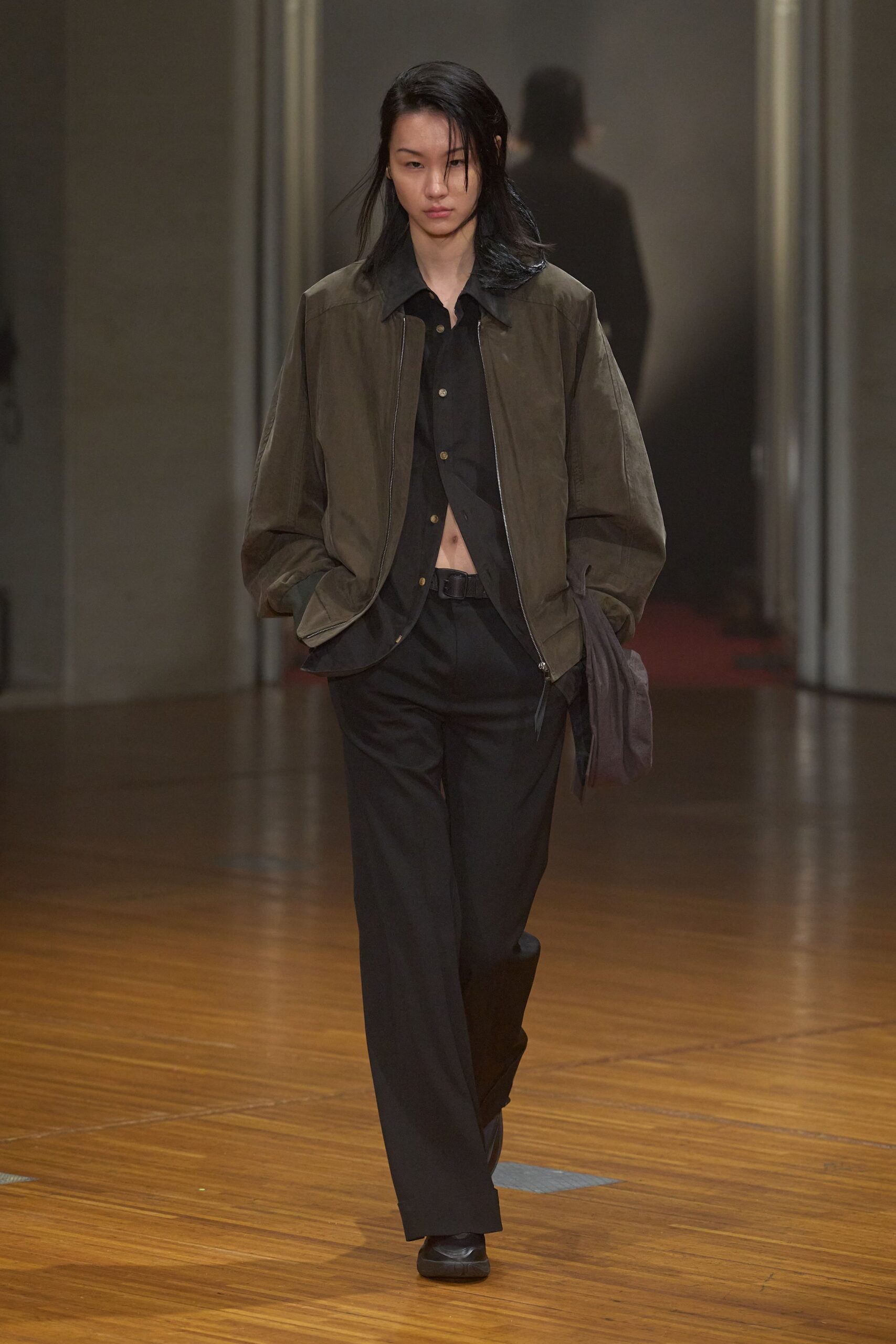 Olive bomber in washed cloth, zip left hanging past the hem. Black button-front shirt, unfastened low to show a narrow strip of stomach. Black straight trousers with a soft break over the shoe. Black belt with a square matte buckle. Square-toe black slip-ons. Walking into the office on Sunday to water a plant you forgot.