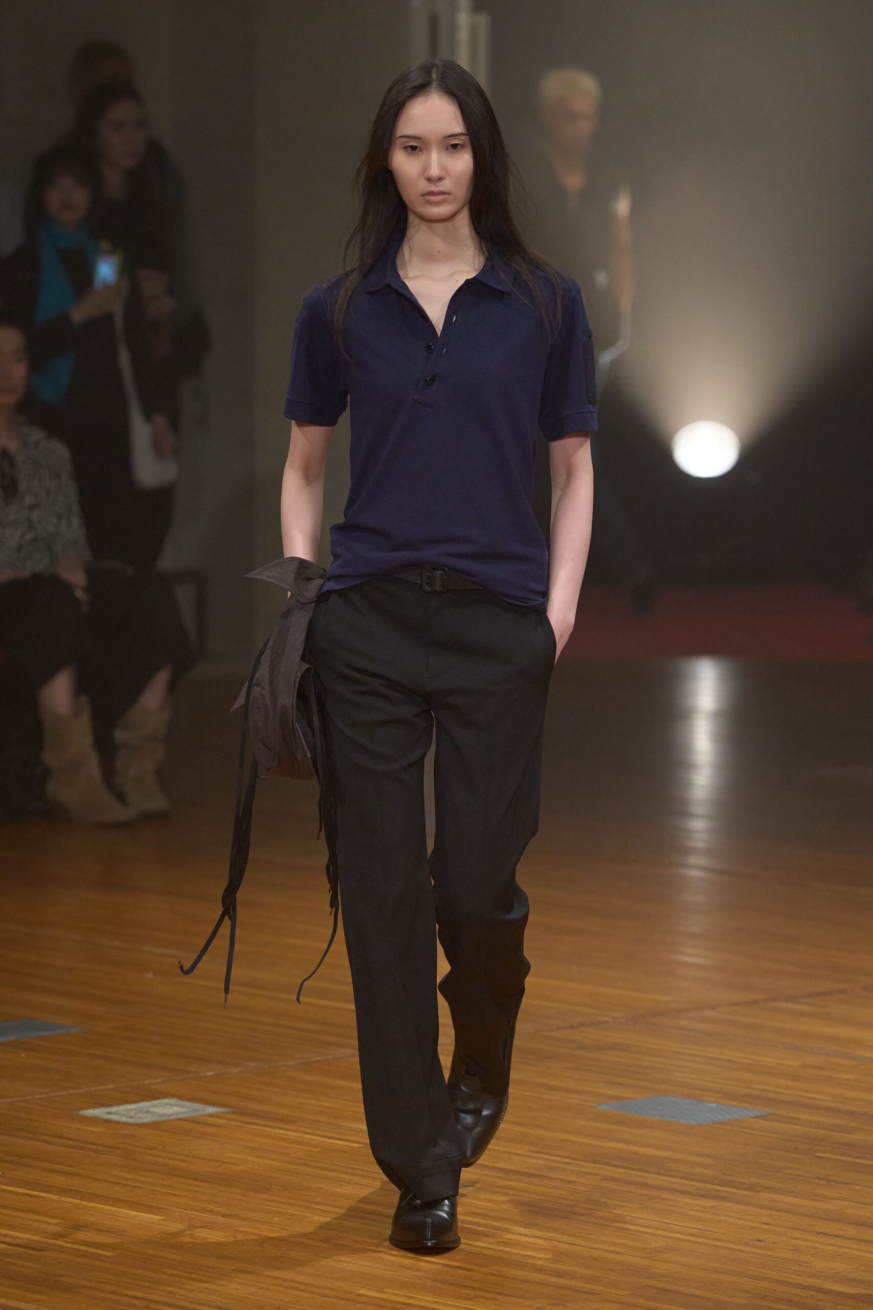 Ink-navy knit polo, placket left open under a soft collar. Black belt with a square buckle. Black straight trousers, hem breaking over the shoe. Black leather square-toe boots. Slouchy black shoulder bag, long straps swinging loose. Walking across an empty civic hall after the chairs have been stacked.