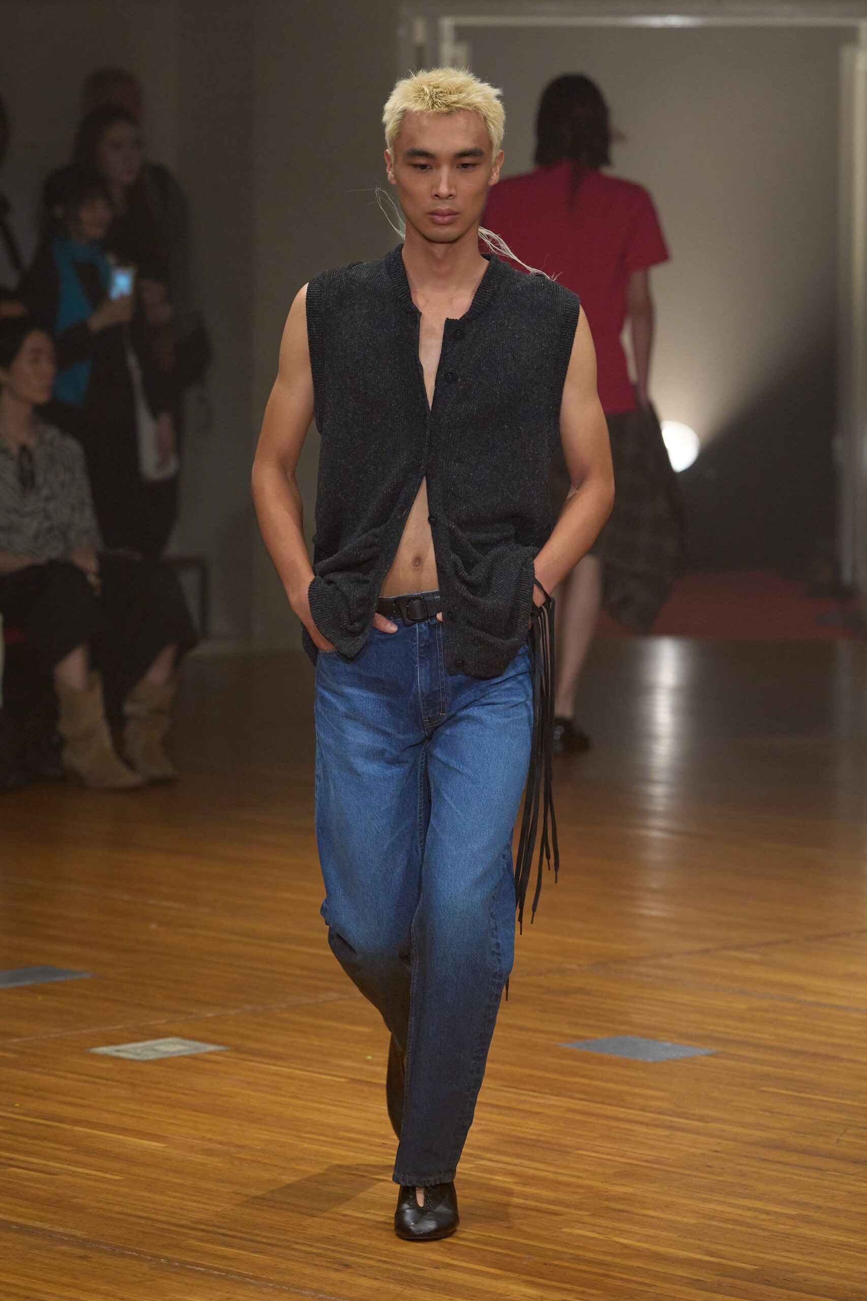 Charcoal ribbed knit hanging open, hem bunching into both pockets. Mid-blue straight jeans, faded softly through the thigh. Black leather belt with long fringe falling off one hip. Black ankle boots with a narrow split opening at the toe. Walking back from the corner store in boots not meant for daylight.
