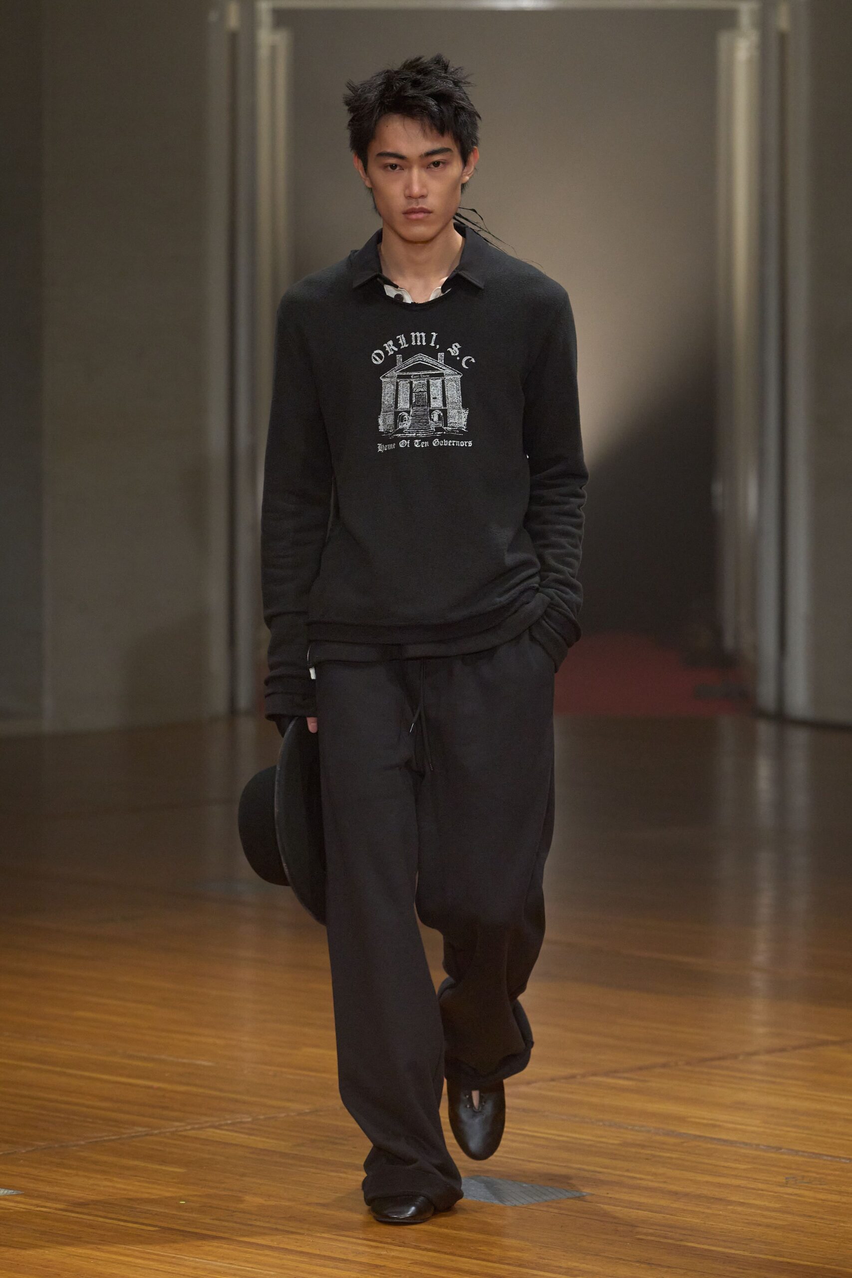 White polo collar peeking unevenly from under the neckline. Black knit sweater with a cracked white building graphic. Black drawstring trousers, cut wide and pooling over the shoes. Black felt hat carried low at the side. Black leather square-toe slip-ons. Whatever you throw on for crossing campus after the library closes.