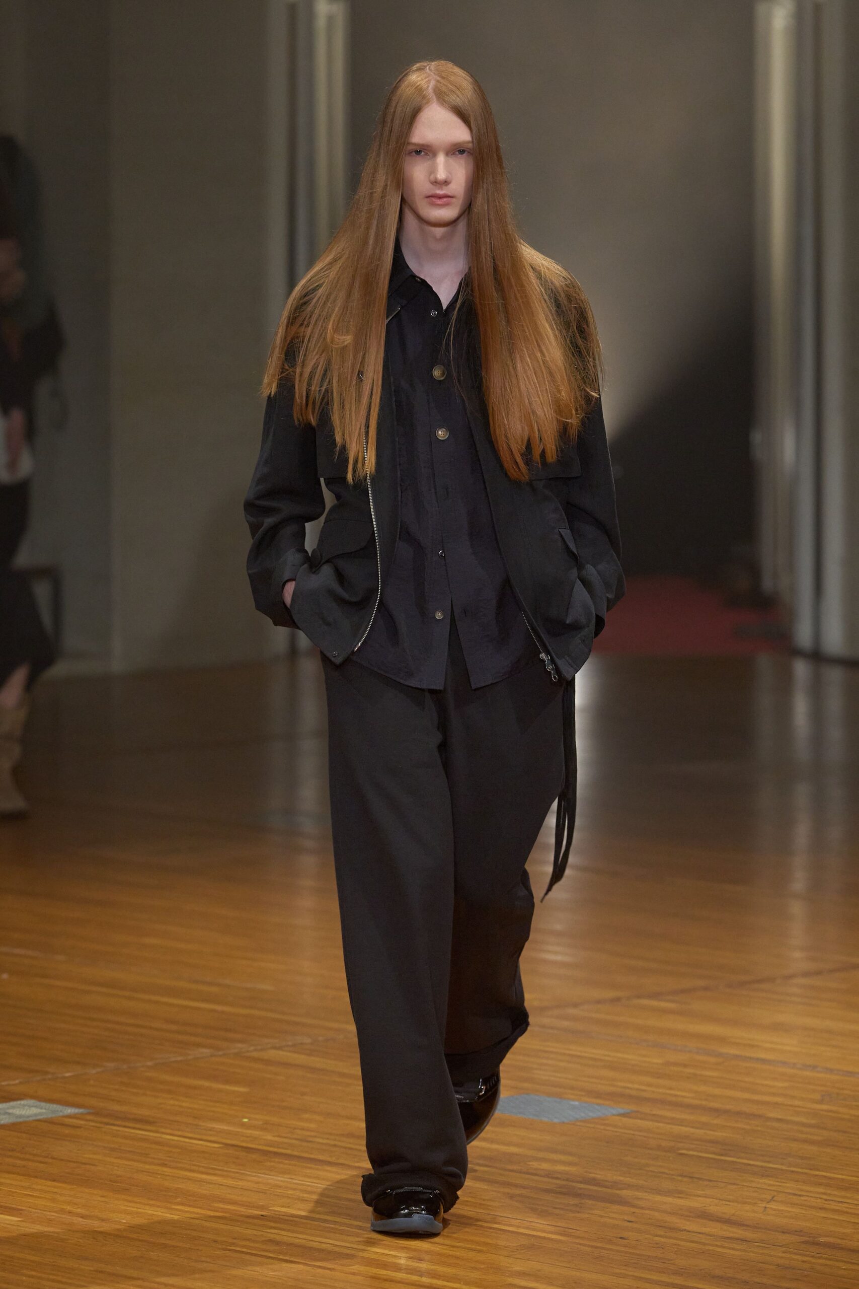 Black zip-front jacket with broad flap pockets and a dangling drawcord end. Washed black shirt, horn-look buttons spaced down the placket. Loose black trousers falling straight with a soft break at the shoe. Glossy black slip-ons with blunt rounded toes. For crossing a civic hall floor before anyone has unlocked the offices.