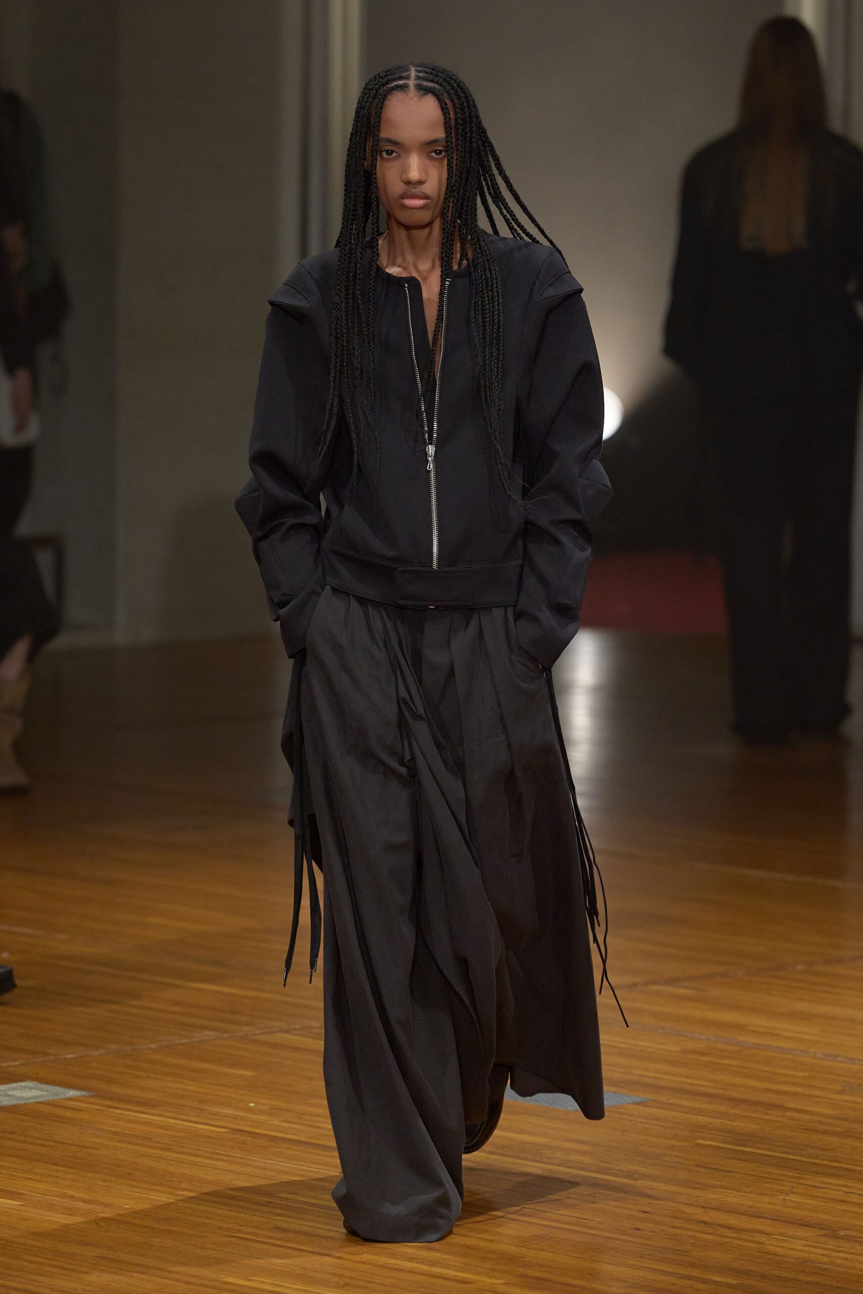 Black zip-front jacket with sculpted shoulder flanges and a low dropped hem. Washed black wide-leg trousers, front pleats collapsing into a wrapped drape. Long black cords hanging loose at each hip. Walking the long hall after rain, keys and receipts lost in your pockets.