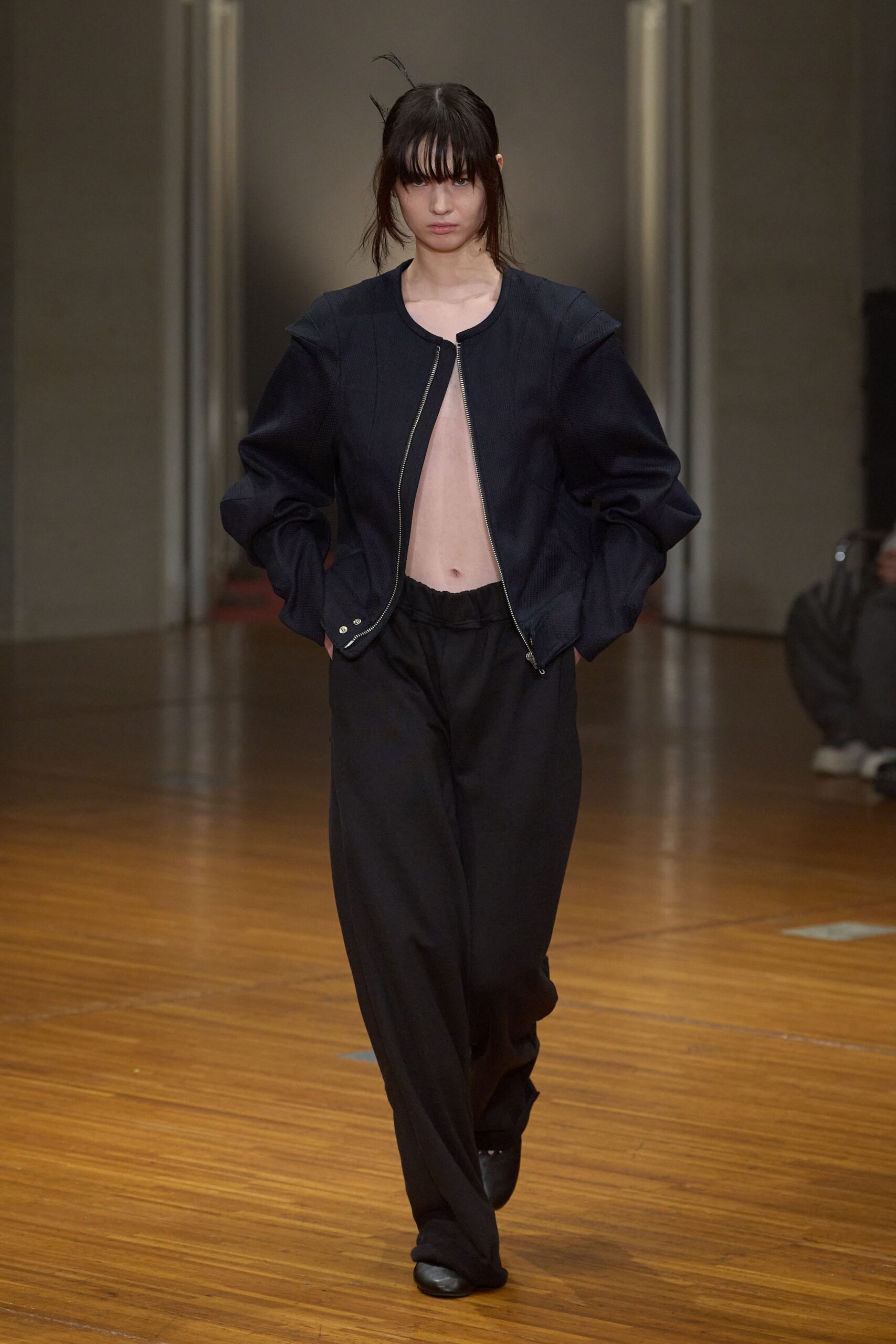 Ink-black textured jacket with sculpted shoulders and an off-center open zip. Loose black trousers with a softly gathered waistband and pooled hem. Square-toe black shoes, nearly swallowed by the trouser break. Crossing a drafty municipal hall after missing the coat check.
