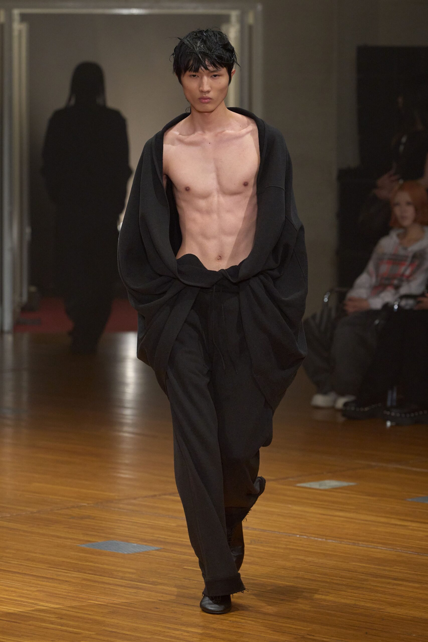 Black knit cardigan hanging off both shoulders, knotted low across the hips. Loose black trousers with a drawstring waist and slightly frayed hems. Square-toe black shoes peeking from the pooled break. Whatever you pull on for dawn crossing a cold, echoing station.