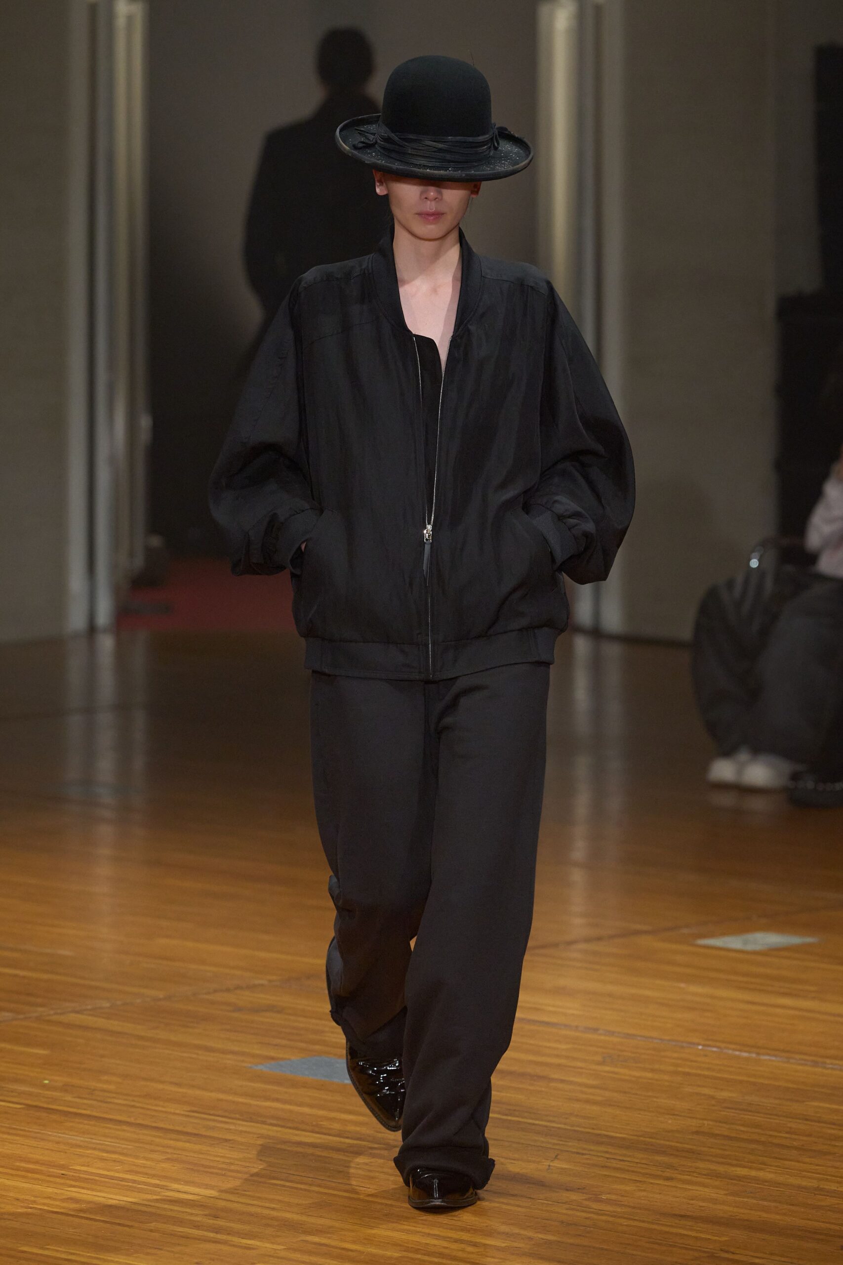 Black felt bowler with a wrapped band and brim tipped over the eyes. Inky lightweight bomber, silver zip ending in a long leather pull. Loose black trousers breaking softly over the shoe. Glossy black square-toe shoes. Walking across a varnished hall floor before anyone unlocks the side doors.