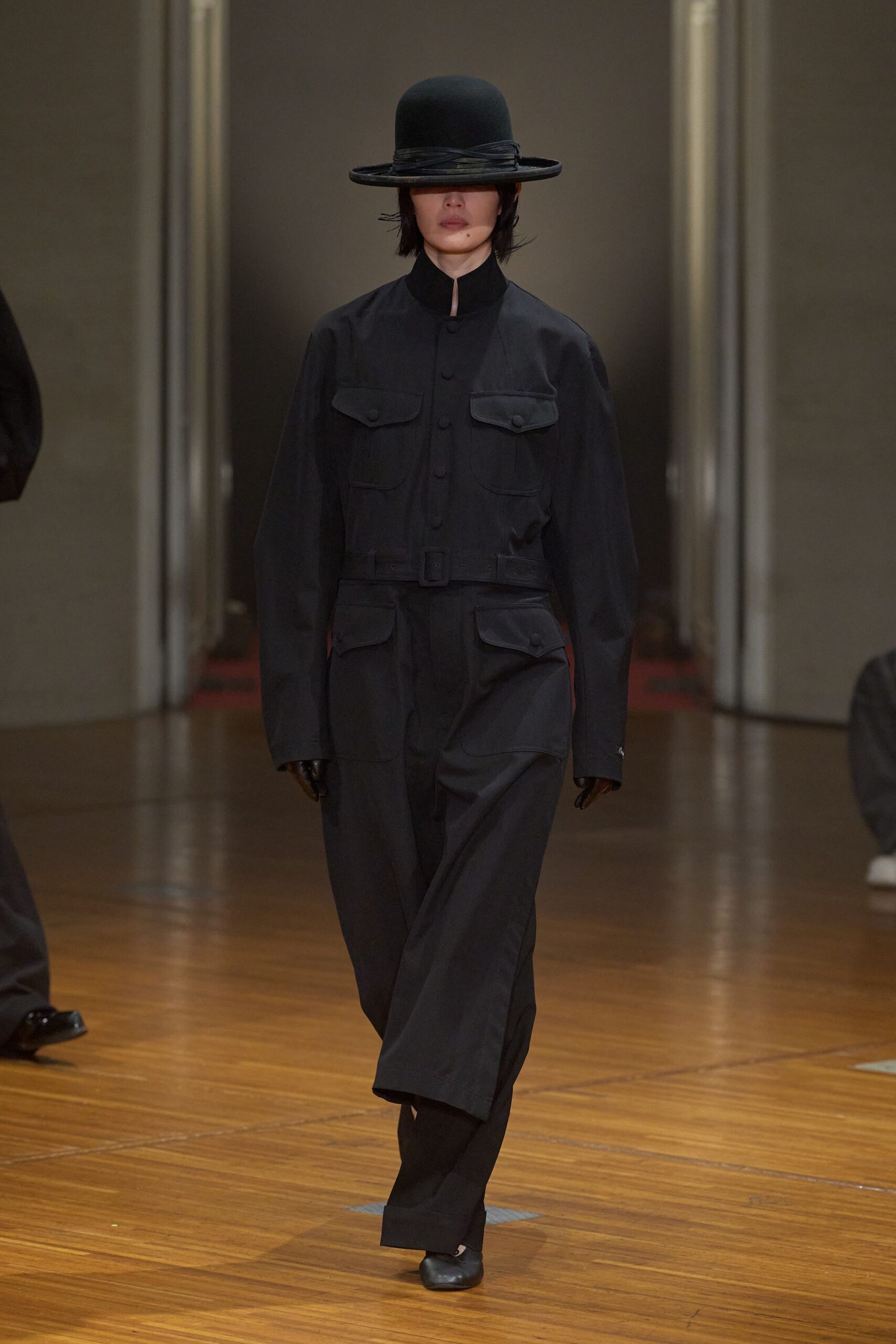 Black bowler hat with a wrapped band and brim shading the eyes. Inky high-collar field jacket, flap pockets and a self-belted waist. Matching straight trousers with a deep break over the shoe. Black leather gloves. Square-toe black shoes. Walking across a civic hall floor before the hearing starts.