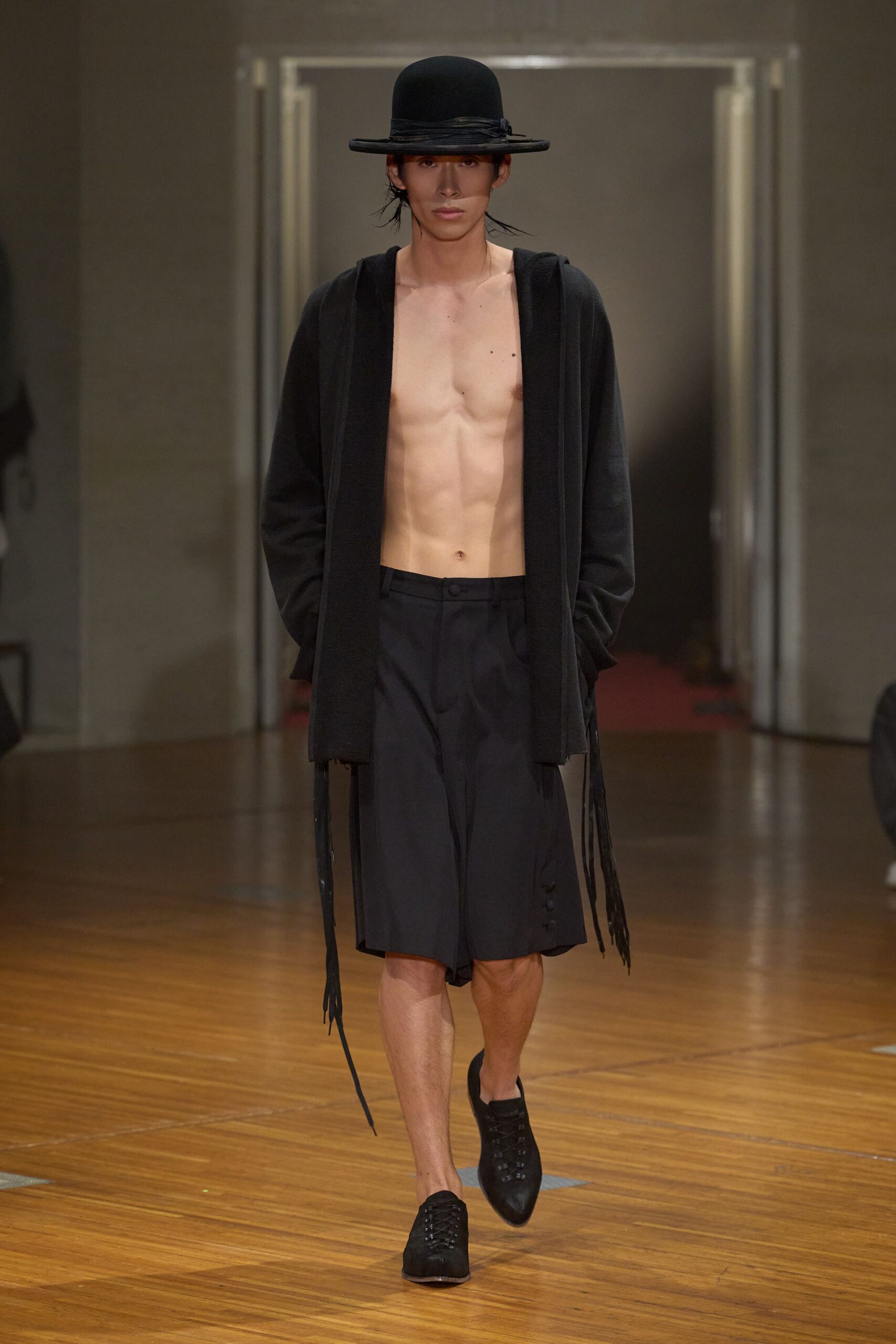 Black bowler with a narrow wrapped band and flat brim. Open black knit cardigan, long leather ties hanging past the hem. High-waisted black tailored shorts, side vent fastened with three buttons. Pointed black lace-ups in matte suede. Crossing a near-empty town hall after a recital let out.