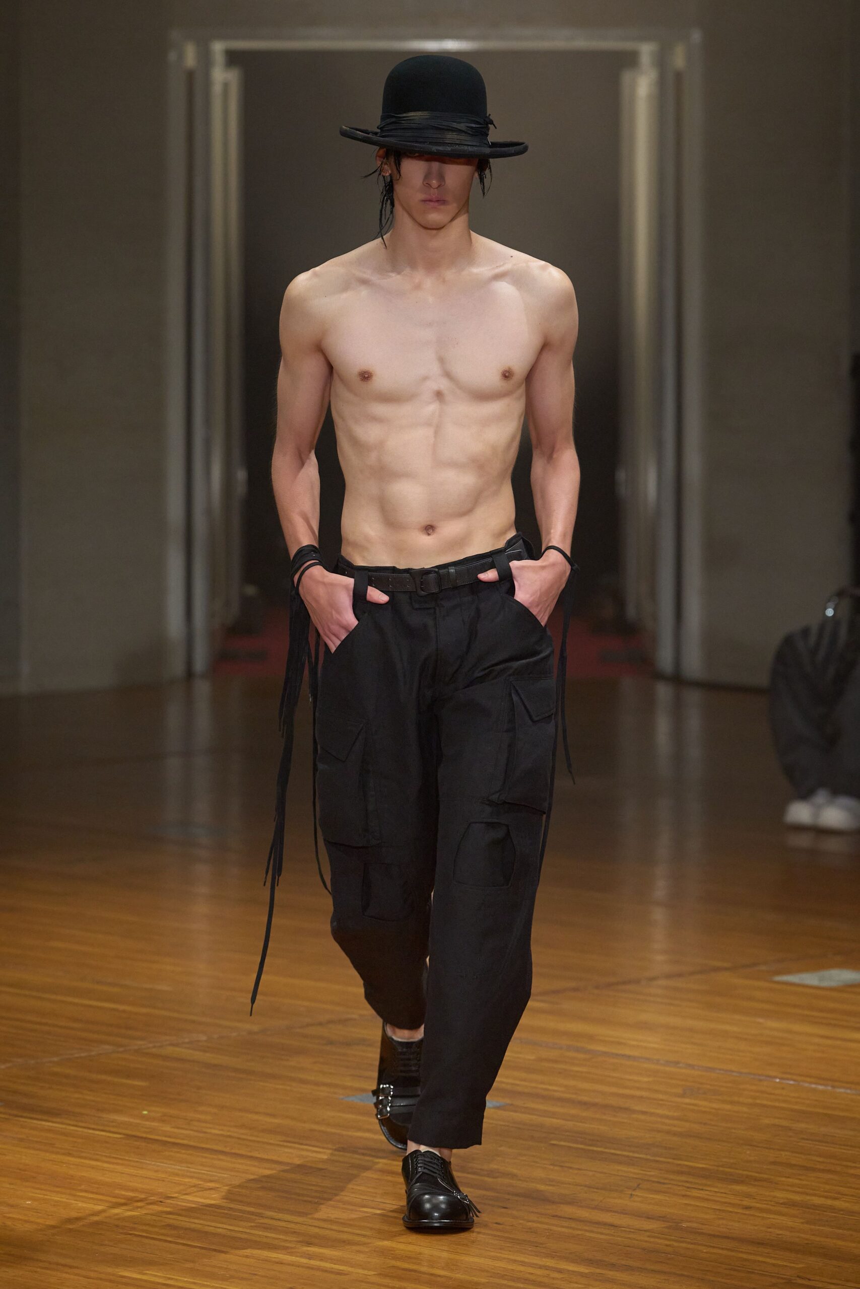 Black felt bowler with a wrapped grosgrain band sinking over one eye. Thin black cords looped close at both wrists. Black cargo trousers with flap pockets and long straps falling from the belt. Black leather lace-ups with fringe hiding the laces. Whatever you wear home when the club smoke is still in your hair.