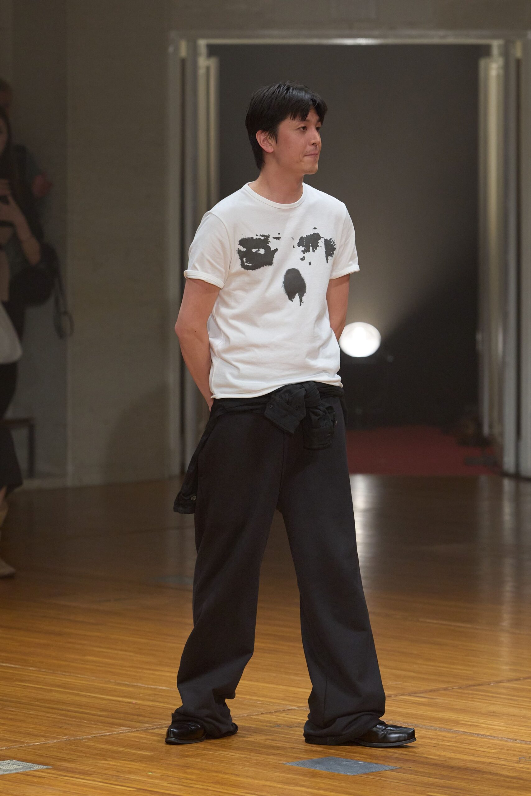 White cotton tee with a distressed black face print, sleeves cuffed once. Black jacket knotted low around the hips, snap tab hanging loose. Loose black trousers breaking heavily over the shoes. Square-toe black leather slip-ons. After-hours in a municipal hall, floor still warm from the afternoon assembly.