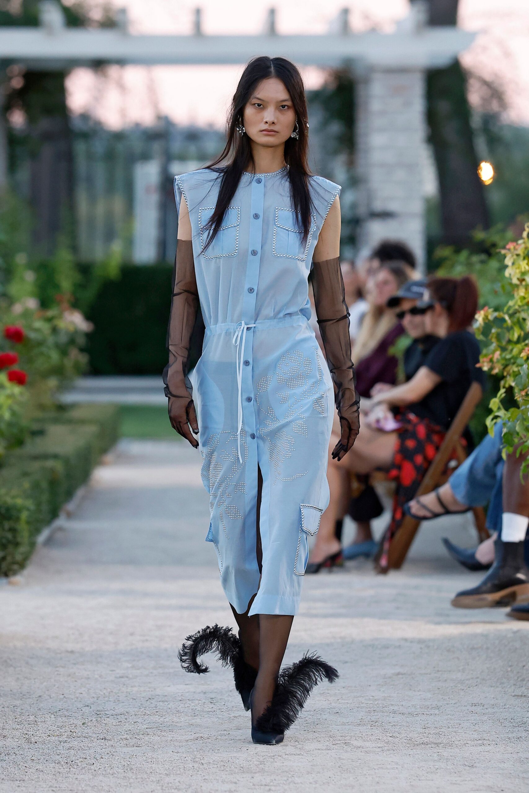 Powder-blue sleeveless shirt dress, crystal-trimmed pockets and a white drawcord waist. Smoke-black mesh opera gloves, wrinkling at the wrists. Black sheer tights. Black pointed boots ringed with long ostrich-feather cuffs. Crystal cluster drop earrings. Rose garden gravel at dusk, after the speeches and before anyone leaves.