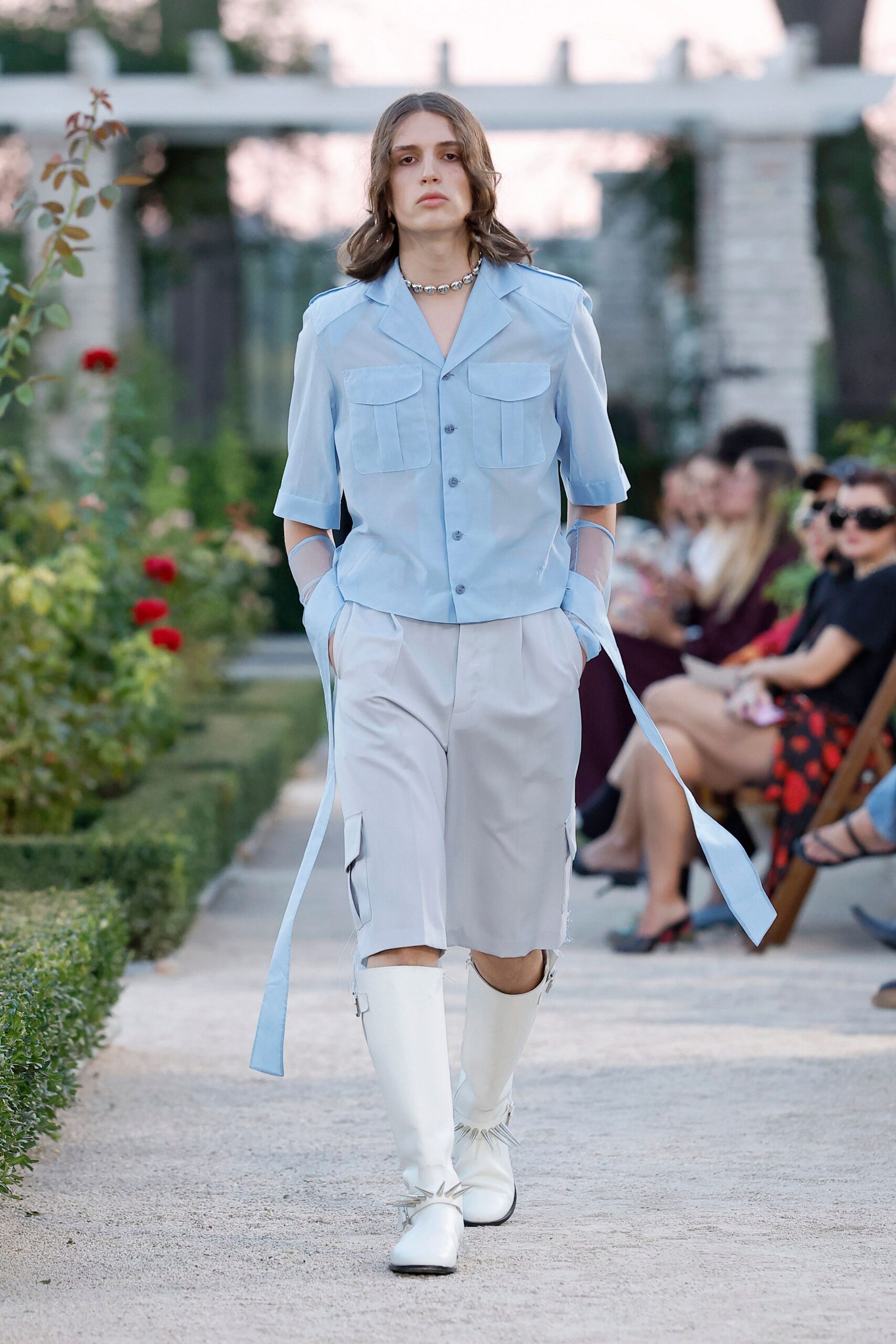 Powder-blue camp shirt with flap pockets and long sash cuffs. Sheer mesh gloves disappearing into the pockets. Pale tailored knee shorts with cargo pockets and raw hems. Chunky silver choker. White leather boots with spur-like metal bursts at the ankle. Walking the rose garden path before dinner is called.