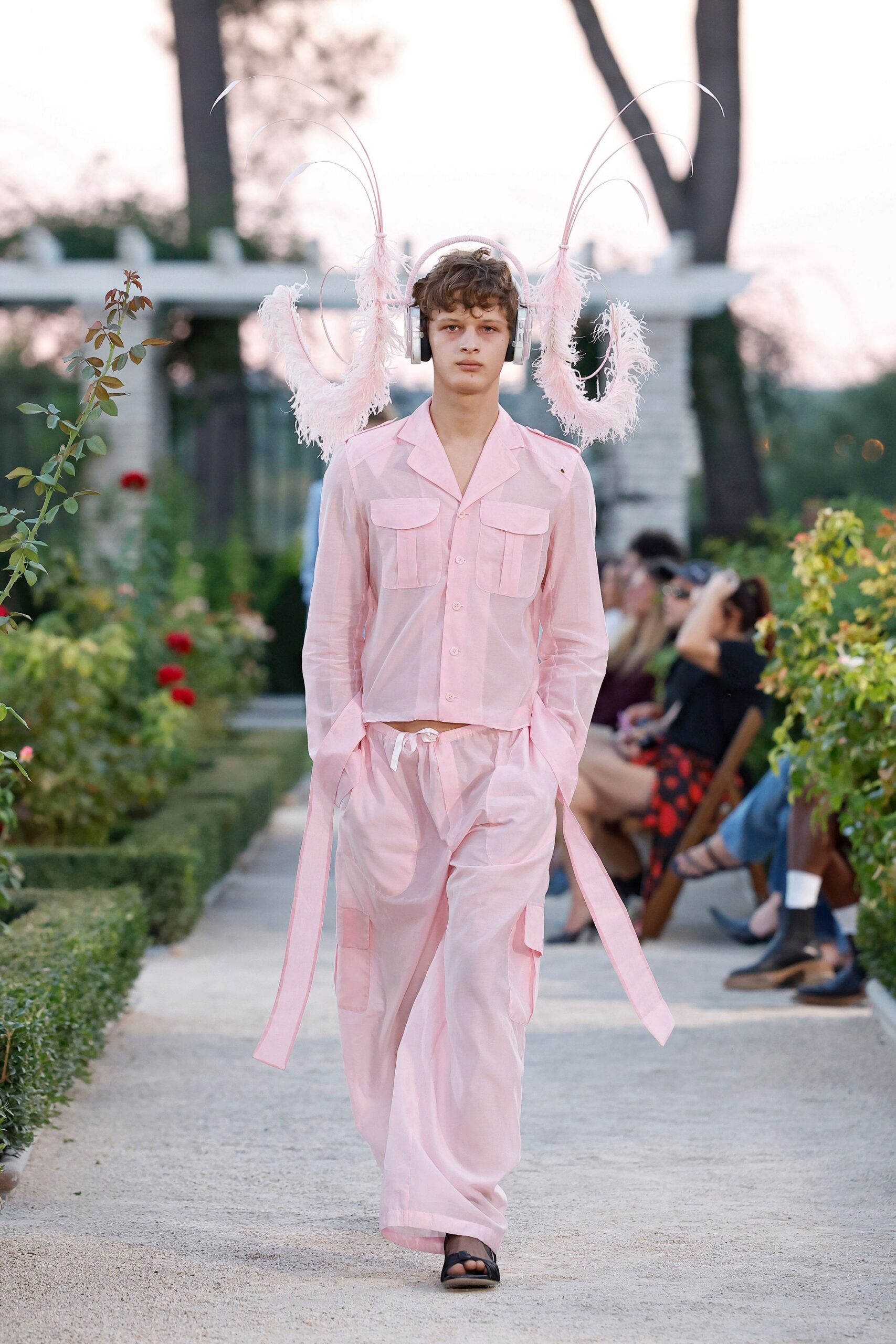 Silver headphones sprouting pale pink feather loops on wiry stalks. Sheer blush camp-collar shirt with twin flap pockets and a cropped hem. Matching drawstring trousers, low slung with oversized cargo pockets and belt ties. Black thong sandals. Walking the rose garden path after someone put the picnic playlist on your head.