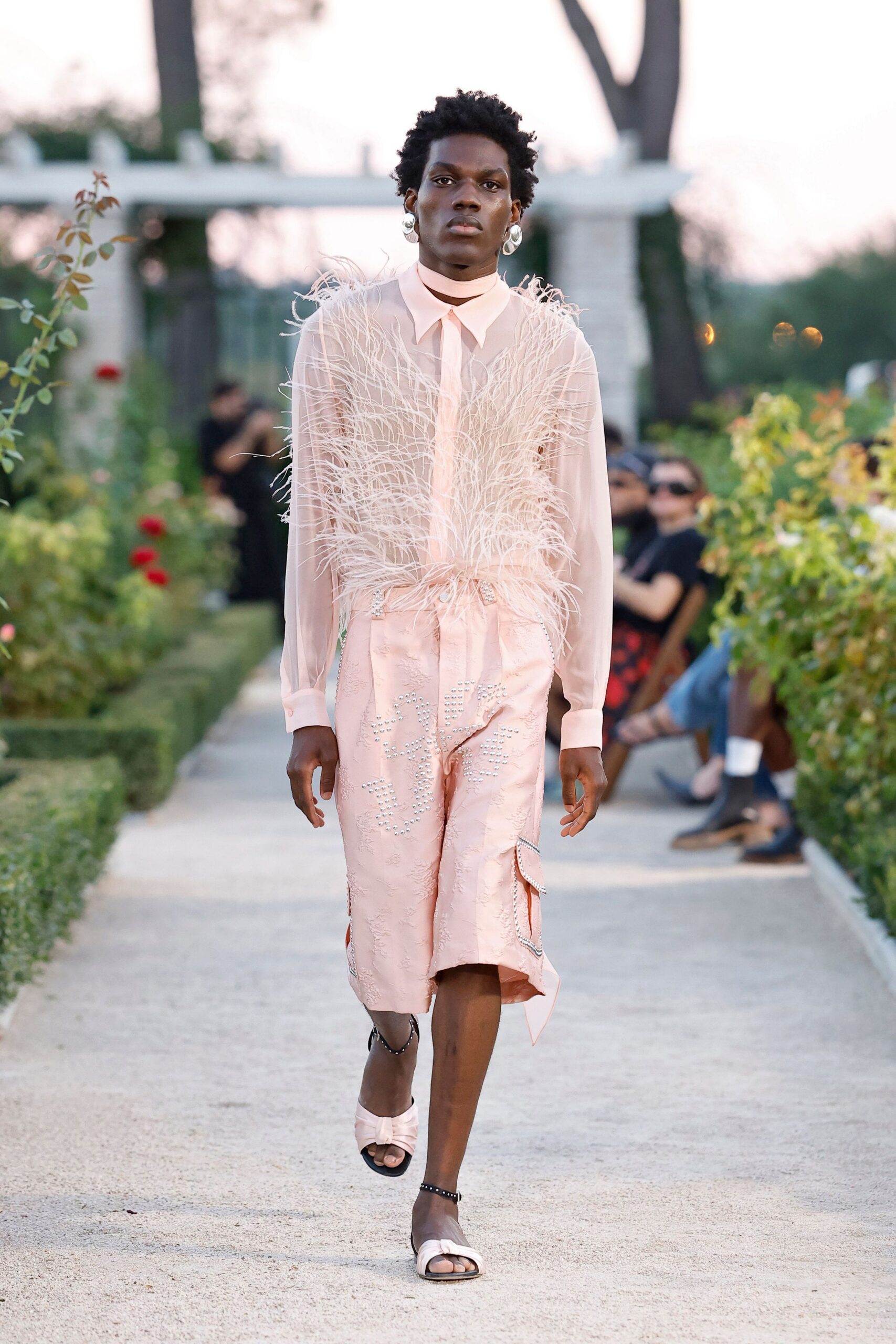 Oversized silver hoops catching the light beside the jaw. Sheer blush blouse with a pointed collar and ostrich-plume front. Blush brocade wrap skirt, crystal studs tracing a curling motif. Pale satin sandals with black studded ankle straps. Rose garden gravel at dusk, when the speeches have finally ended.