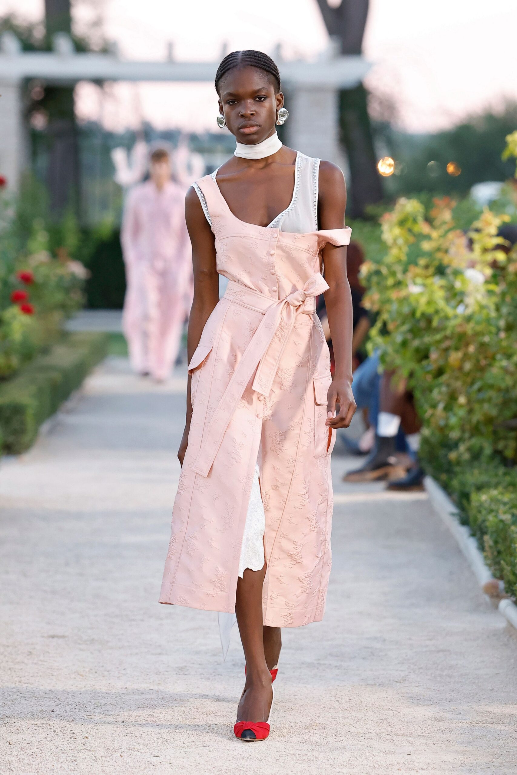 Wide white band of smooth satin wrapped close at the throat. Pearled straps and a scooped neckline on sheer white underlay. Blush floral jacquard falling off one shoulder, sash knotted long. Crumpled silver discs. White pumps with red bow toes. Rose garden aisle just before the string quartet starts.