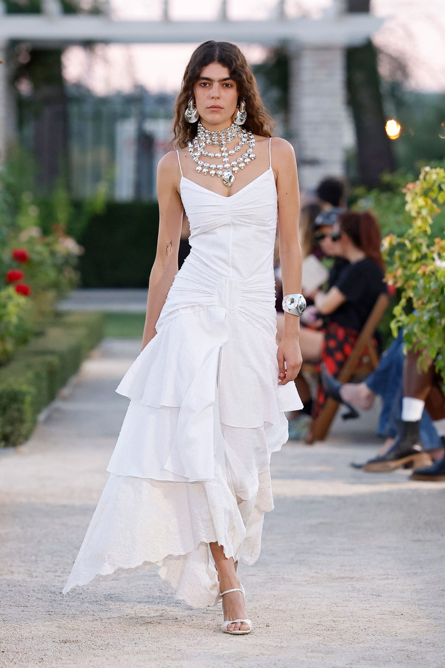 Layered silver medallion chokers and chains, one pendant hanging heavy at the sternum. Oversized hammered silver hoops. White slip dress in ruched dotted chiffon, dropped into uneven tiered flounces. Wide mirror-bright silver cuff. White ankle-strap sandals. Garden wedding after the vows, gravel stuck to the hem.