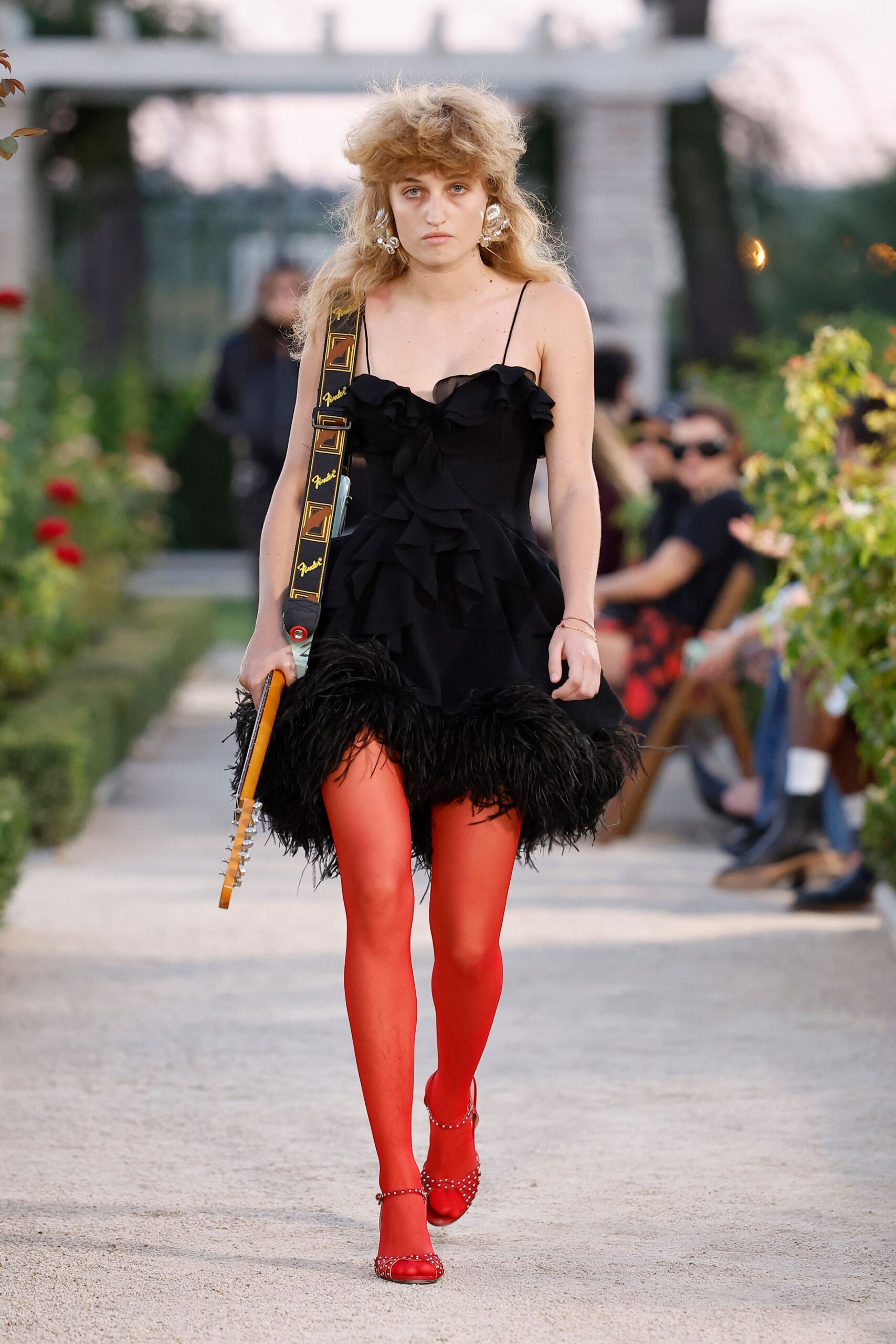 Black slip dress with ruffled tiers and a shaggy feather hem. Tomato-red tights, opaque and startling under the black. Red satin Mary Janes ringed with silver studs. Electric guitar slung on a logo strap, neck hanging by her knee. Crumpled silver disc earrings. Crossing the vineyard gravel just before the first song starts.