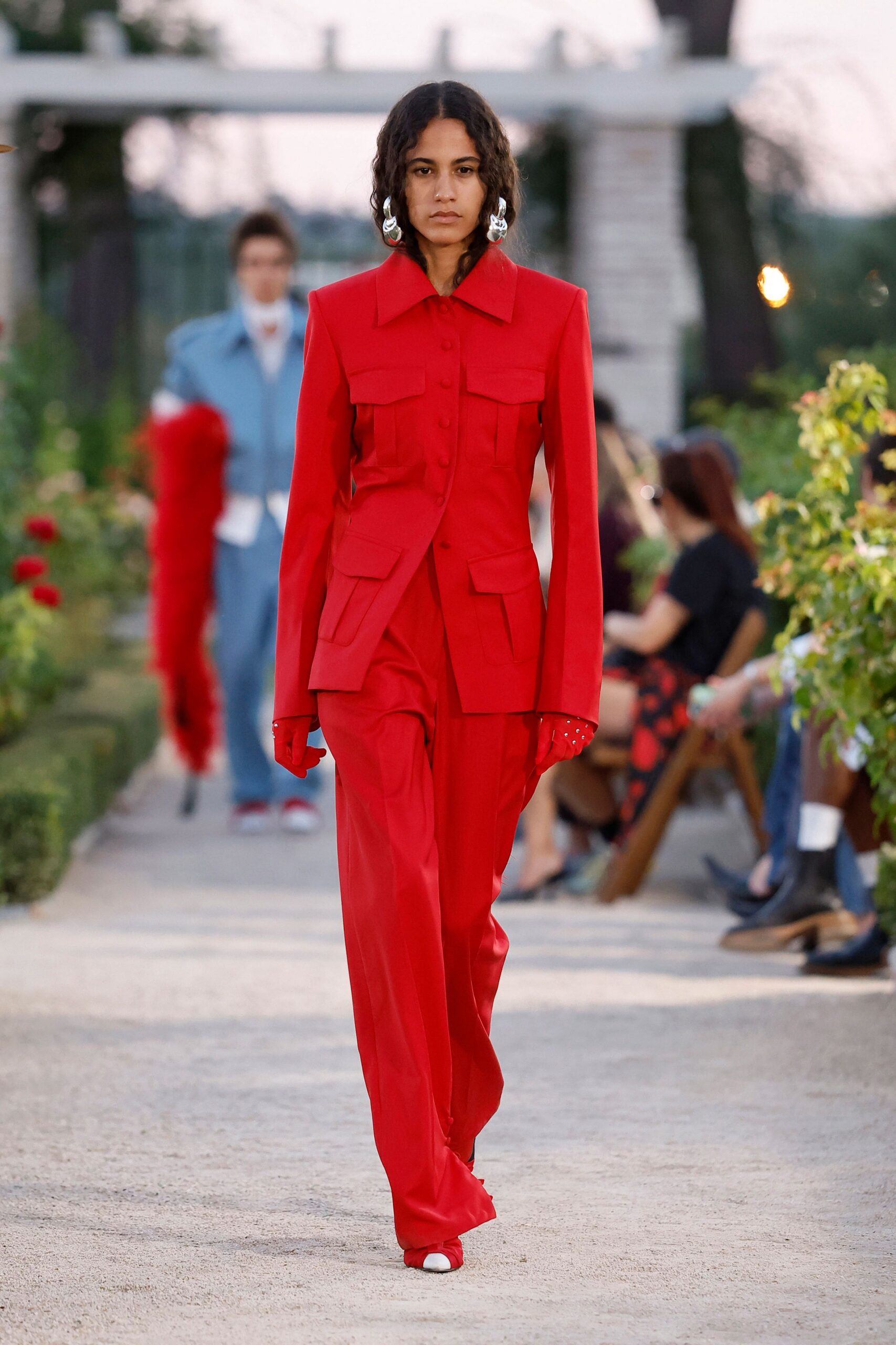 Oversized molten silver hoops brushing the jaw. Scarlet tailored jacket with broad collar and four flap pockets. Matching gloves, silver studs pricking the cuff edge. Fluid scarlet trousers breaking long over the shoe. White square toes peeking from the hem. Crossing a gravel courtyard just before the wedding guests sit down.