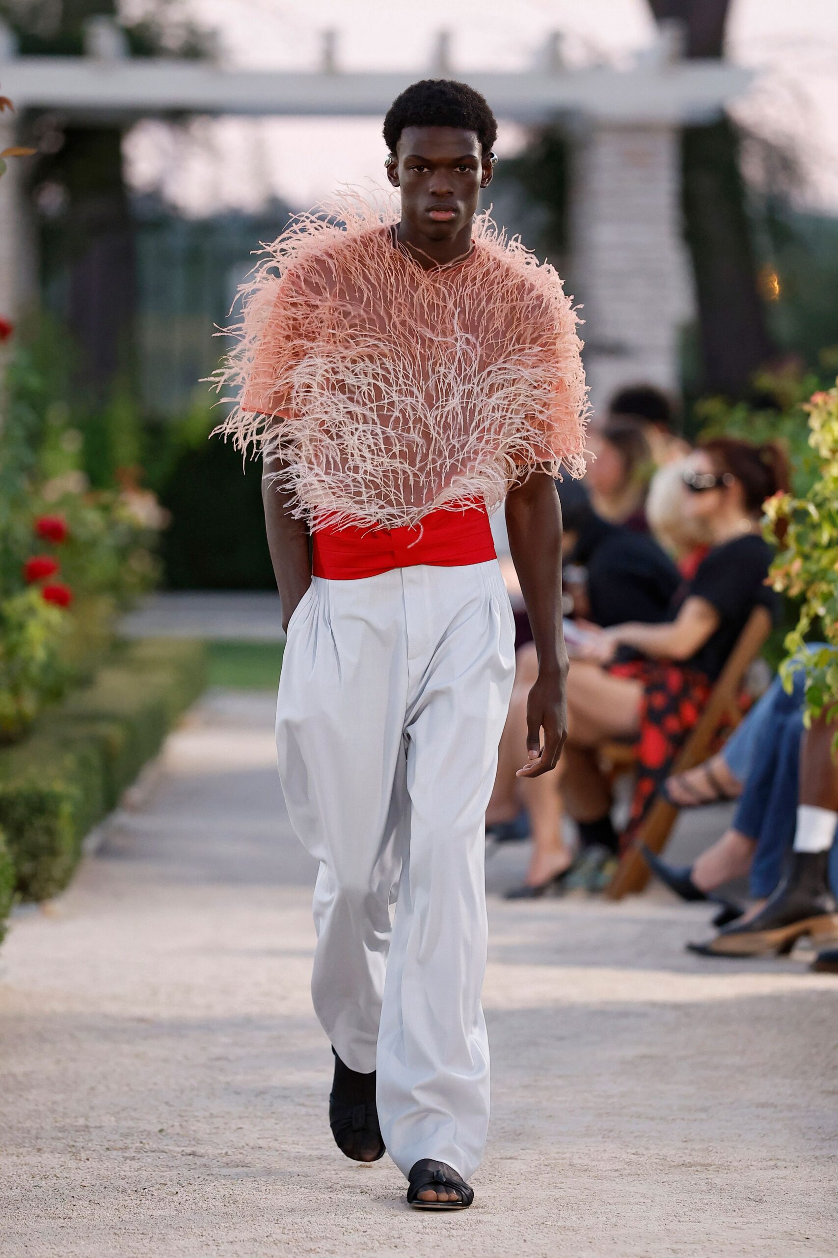 Peach sheer mesh top buried under dense ostrich-frond trim. Wide scarlet cummerbund wrapped flat across the waist. Ivory wide-leg trousers with deep pleats falling from the waistband. Black leather toe-loop sandals. Garden path at dusk, roses throwing scent harder than the heat.