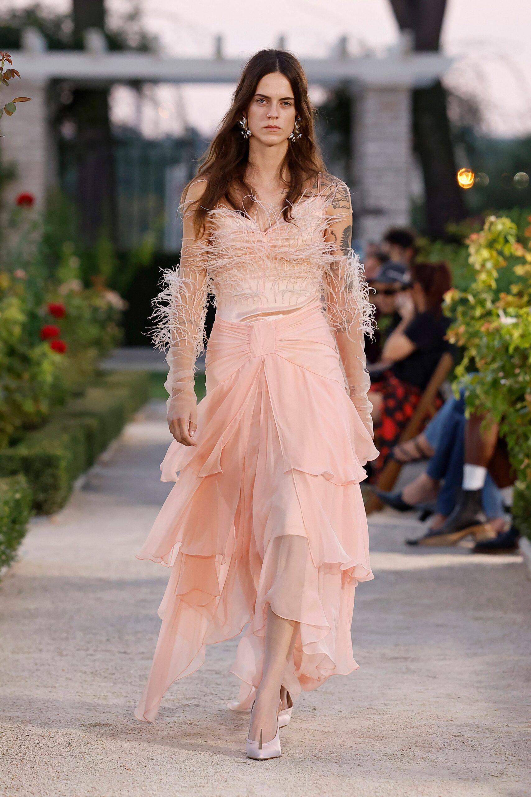 Blush chiffon gown with ostrich-feather bodice and a gathered knot at the waist. Sheer nude tulle sleeves, feathered from elbow to wrist. Pale satin pumps with squared vamps. Silver floral drop earrings. Garden wedding after sunset, gravel catching in the hem.
