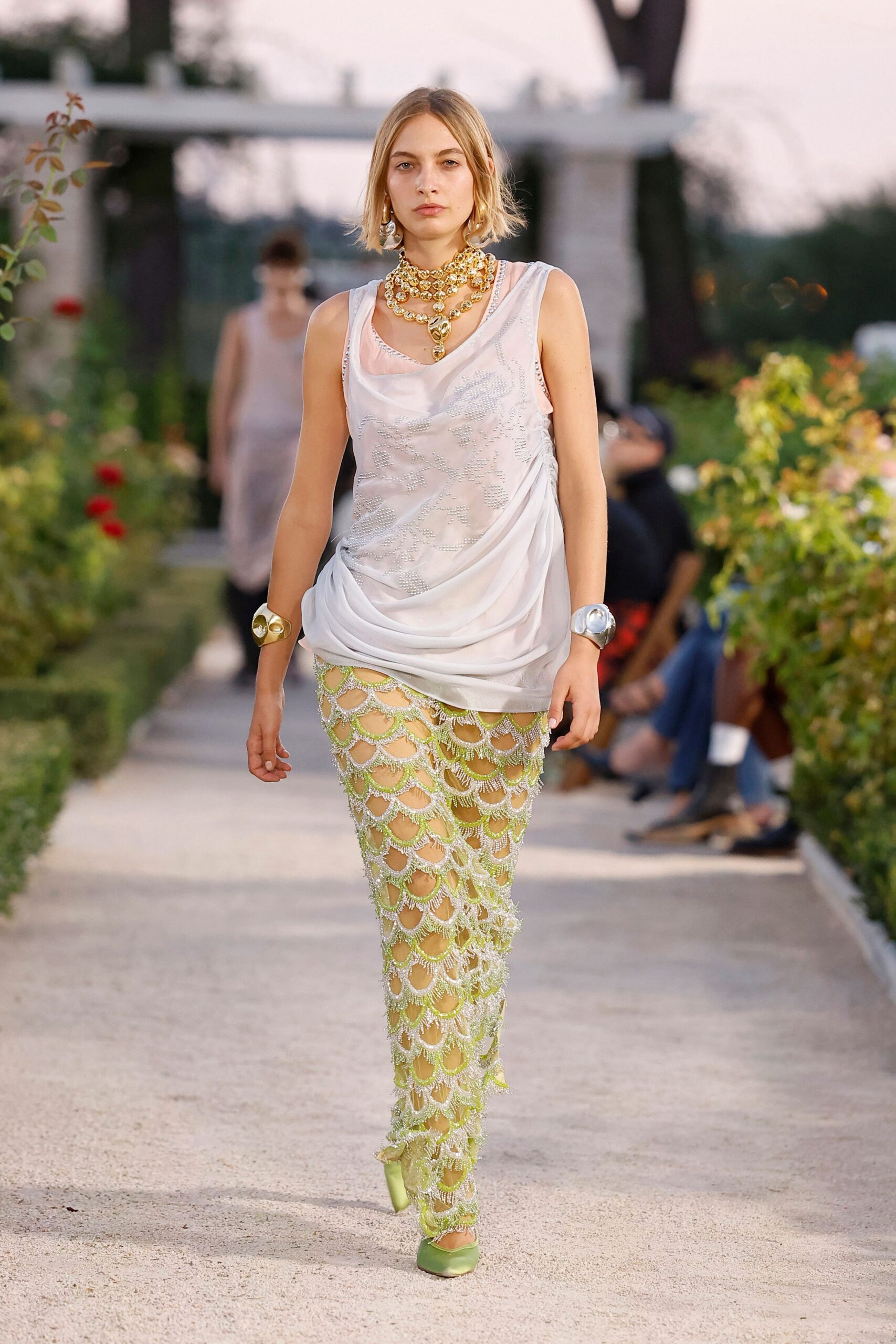 Tiered gold bead chokers with a bulbous drop pendant. Blush sheer draped tank, silver beadwork ghosting across the front. Lime net trousers crusted with scalloped crystal fringe. Pale green satin pumps. Mismatched metal cuffs, one gold one silver. Garden path at dusk, somebody has just lit the citronella candles.