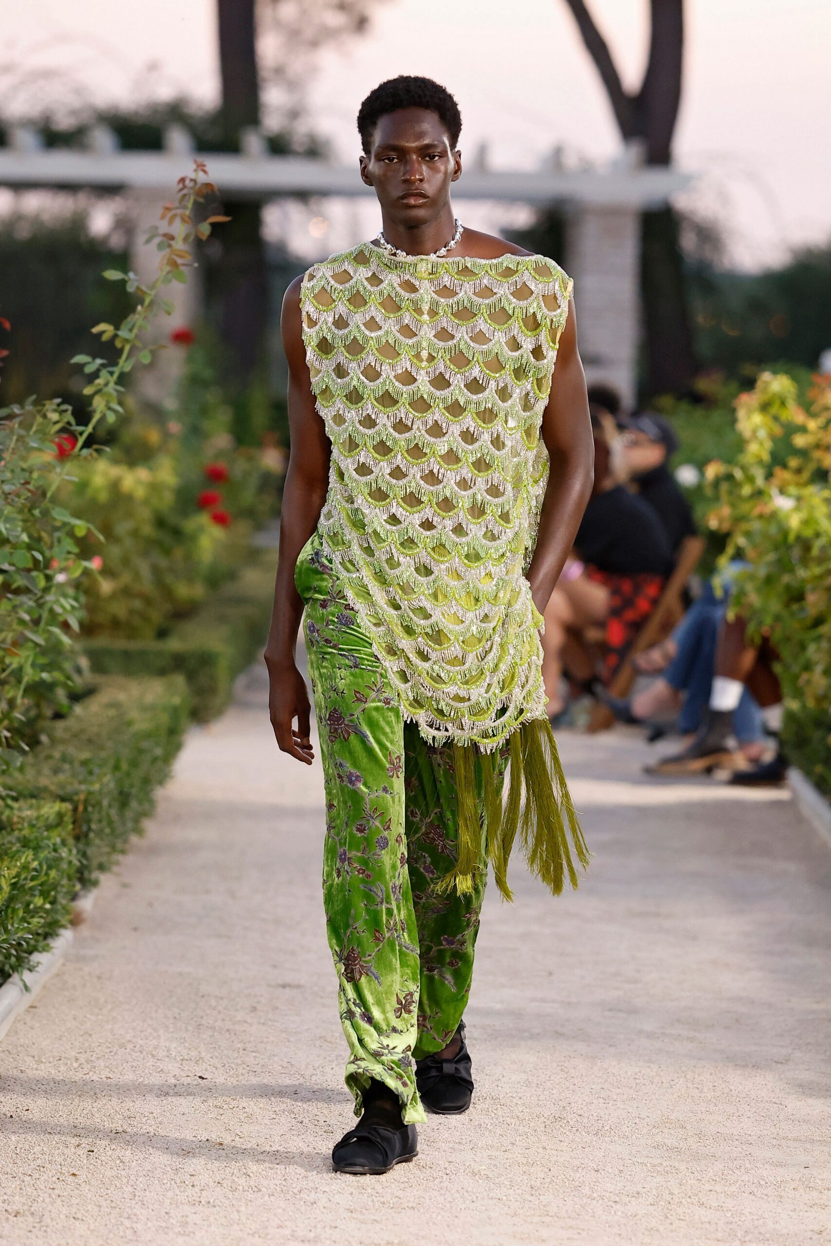 Chunky silver links tight at the collarbone. Sleeveless beaded net tunic in scalloped rows, green tassels dragging at one hip. Chartreuse devore velvet trousers scattered with dusky floral stems. Black satin slip-ons, softly ruched over the foot. Crossing a walled garden after rain, gravel still damp underfoot.
