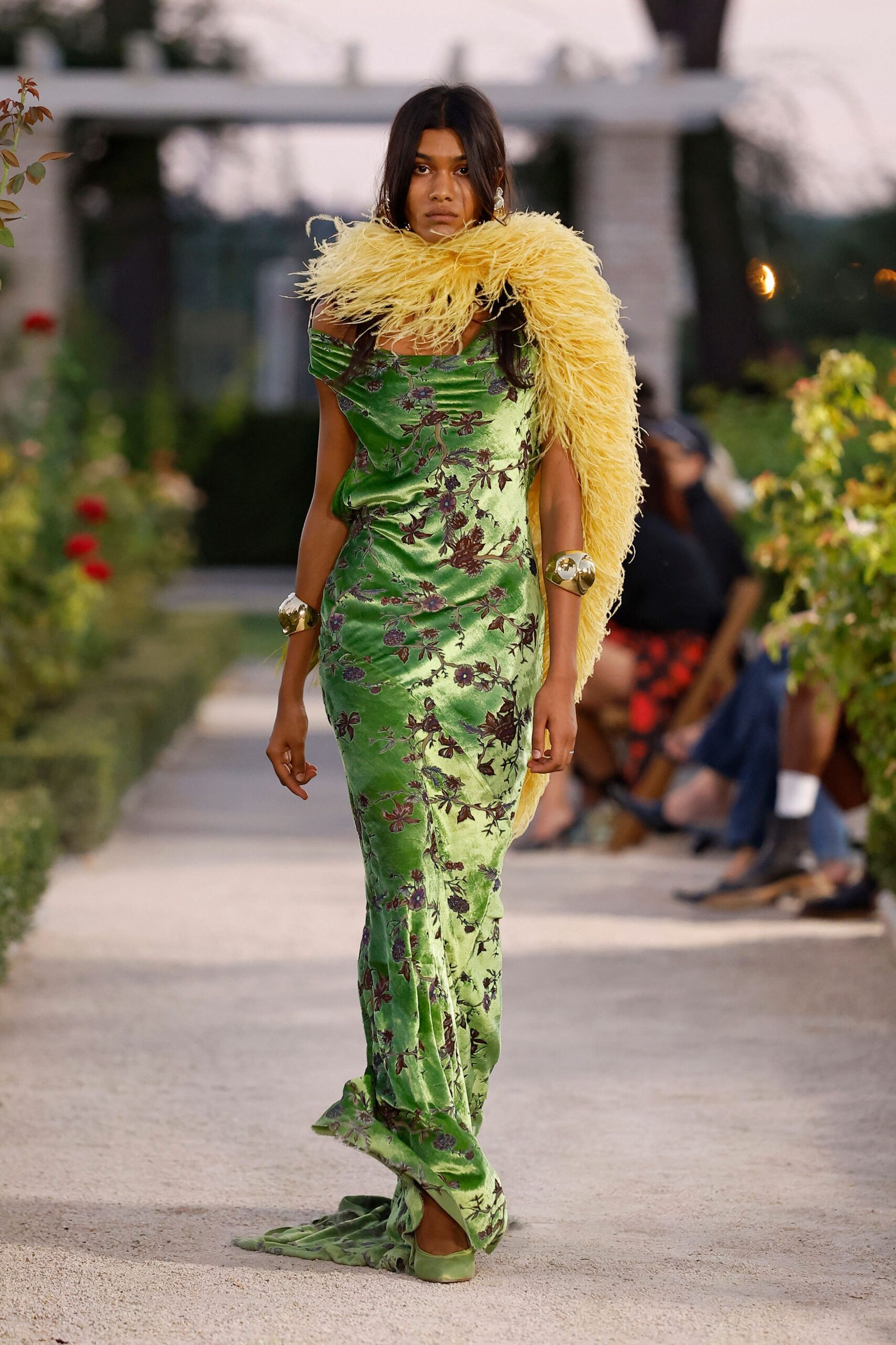 Butter-yellow ostrich feather stole looped huge around the shoulders. Apple-green bias-cut satin gown with shadowy floral brocade and a pooling hem. Mirror-bright gold cuffs, one perched high above the elbow. Green satin flats, square at the toe. Crossing a villa garden at dusk while the champagne is still being poured.