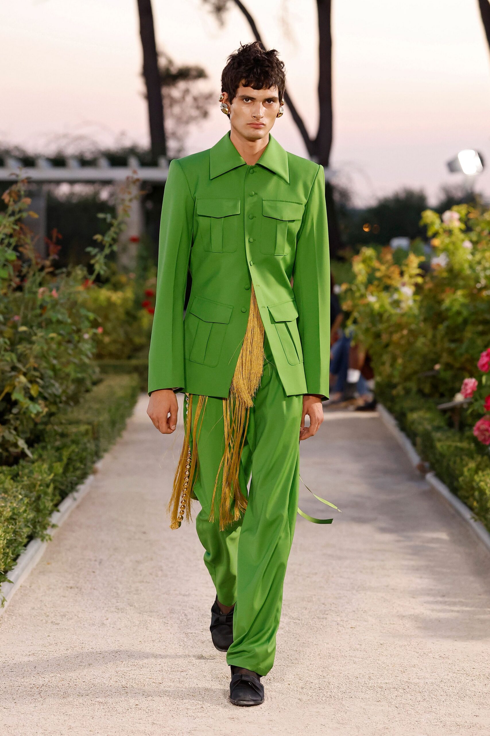 Acid-green tailored jacket with exaggerated collar and four flap pockets. Long straw fringe spilling from the half-buttoned front. Matching fluid trousers, a crystal-trim strip glinting down one side. Square black shoes. Crossing a formal garden at dusk with raffle tickets in your pocket.