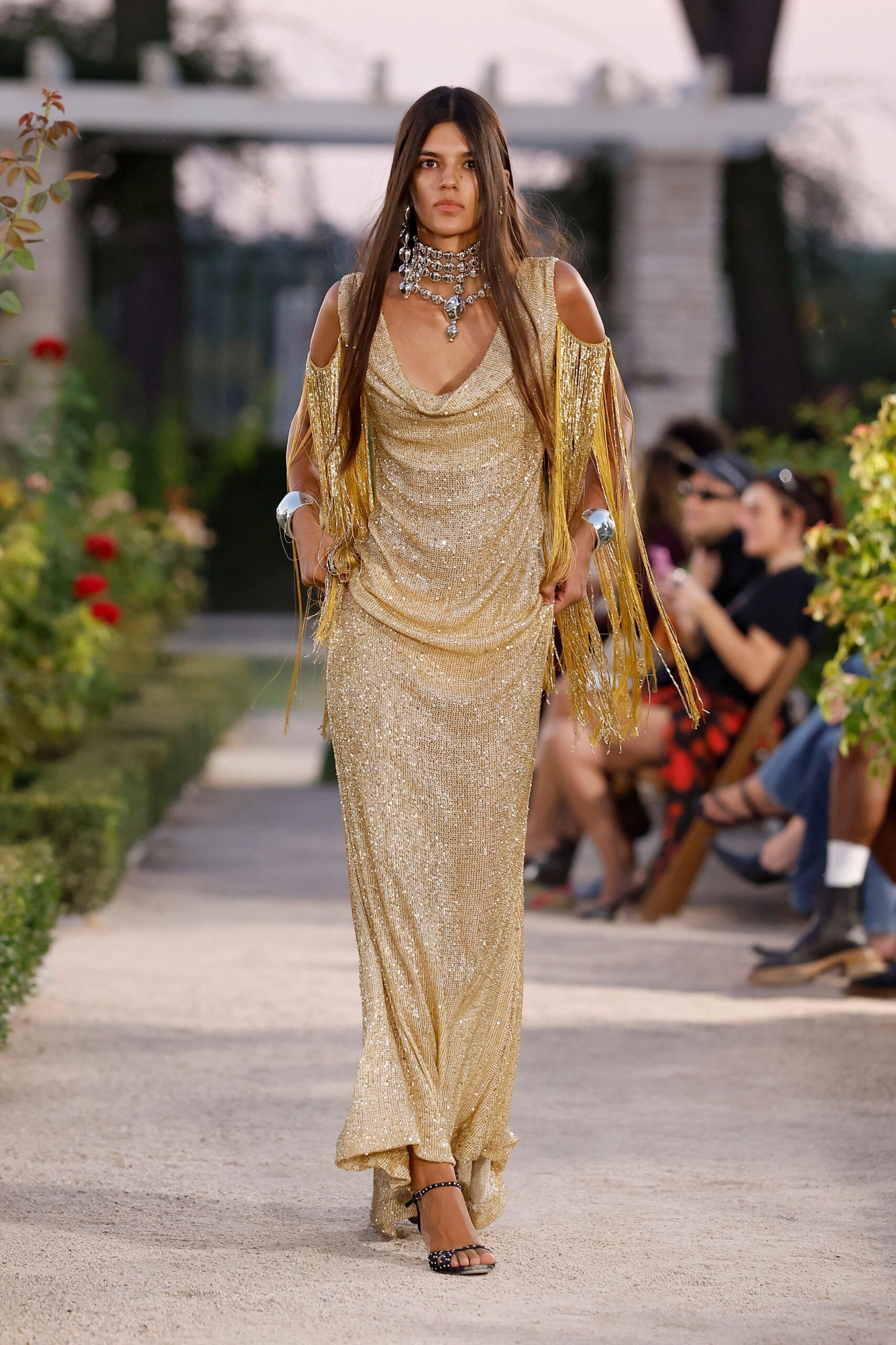 Stacked silver chokers crowded with teardrop pendants. Gold sequin column gown with cowl neck and shoulder cutouts. Long fringe panels swinging from the upper arms. Mirror-bright silver cuffs at both wrists. Black studded ankle-strap sandals. Garden party at dusk, someone has dragged the speakers onto the lawn.