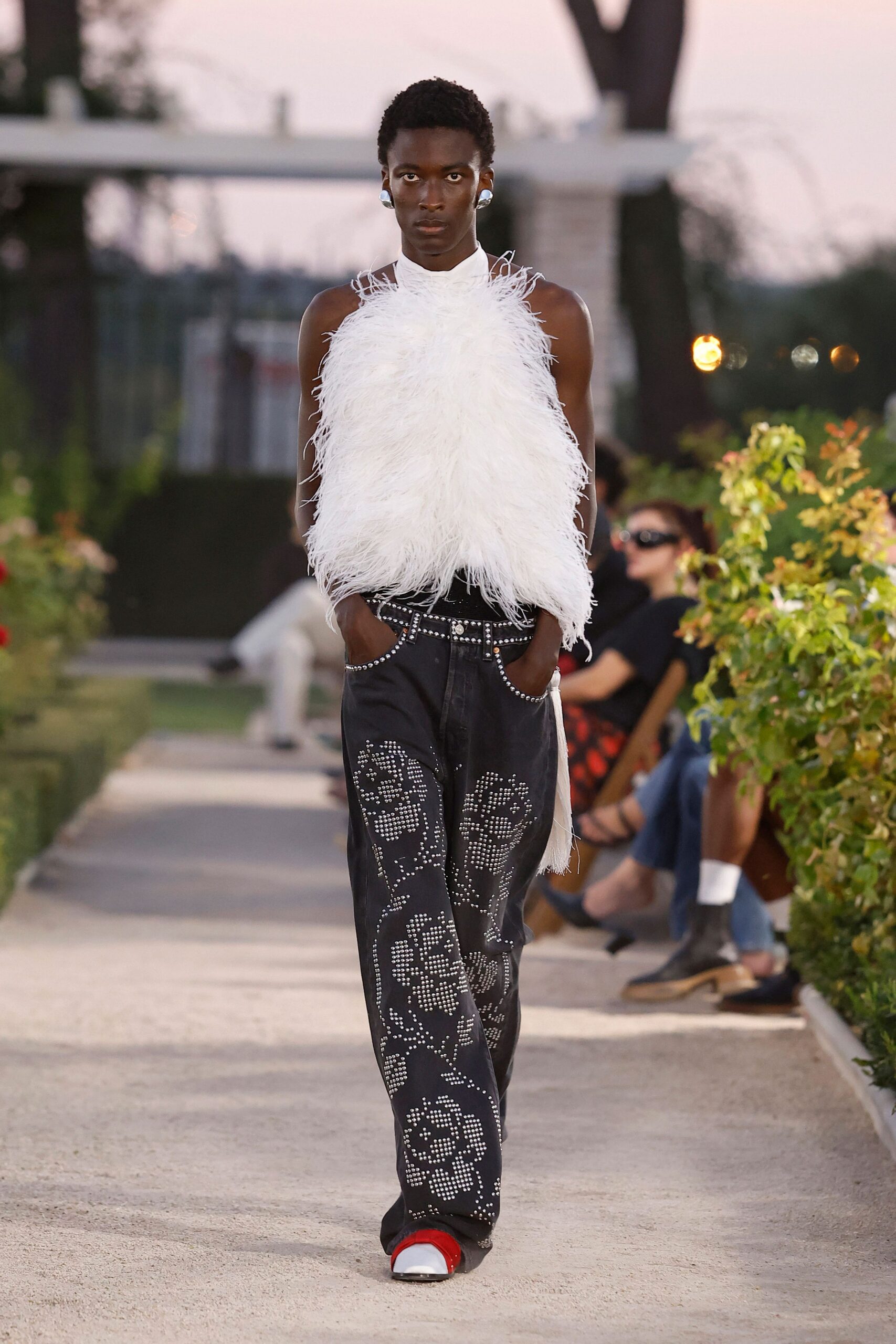 Bulbous silver clip earrings catching the last light. White halter top buried under long ostrich-feather fringe. Black jeans studded with scrolling floral motifs and crystal-rimmed pockets. Square-toe shoes with a flash of red at the vamp. Garden party winding down, someone has started picking roses in the dark.